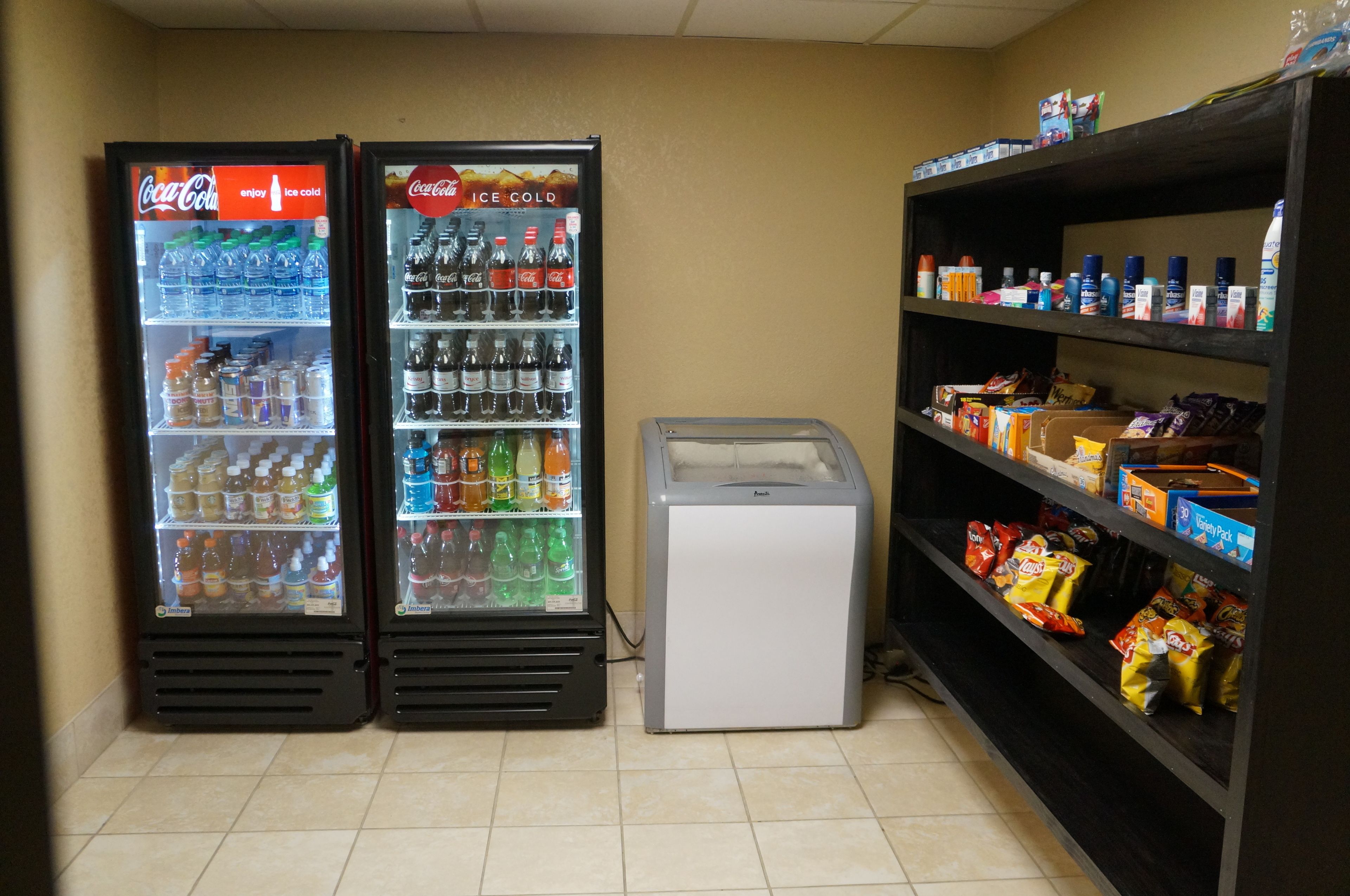 Two Coca-Cola refrigerators stocked with beverages, a small freezer, and shelves with snacks and toiletries.