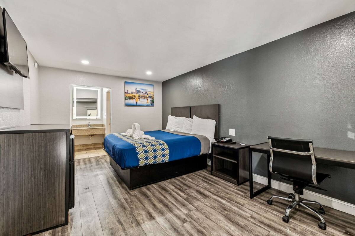 A hotel room with a bed, a desk, a chair, a TV, a mirror, and a painting on the wall.