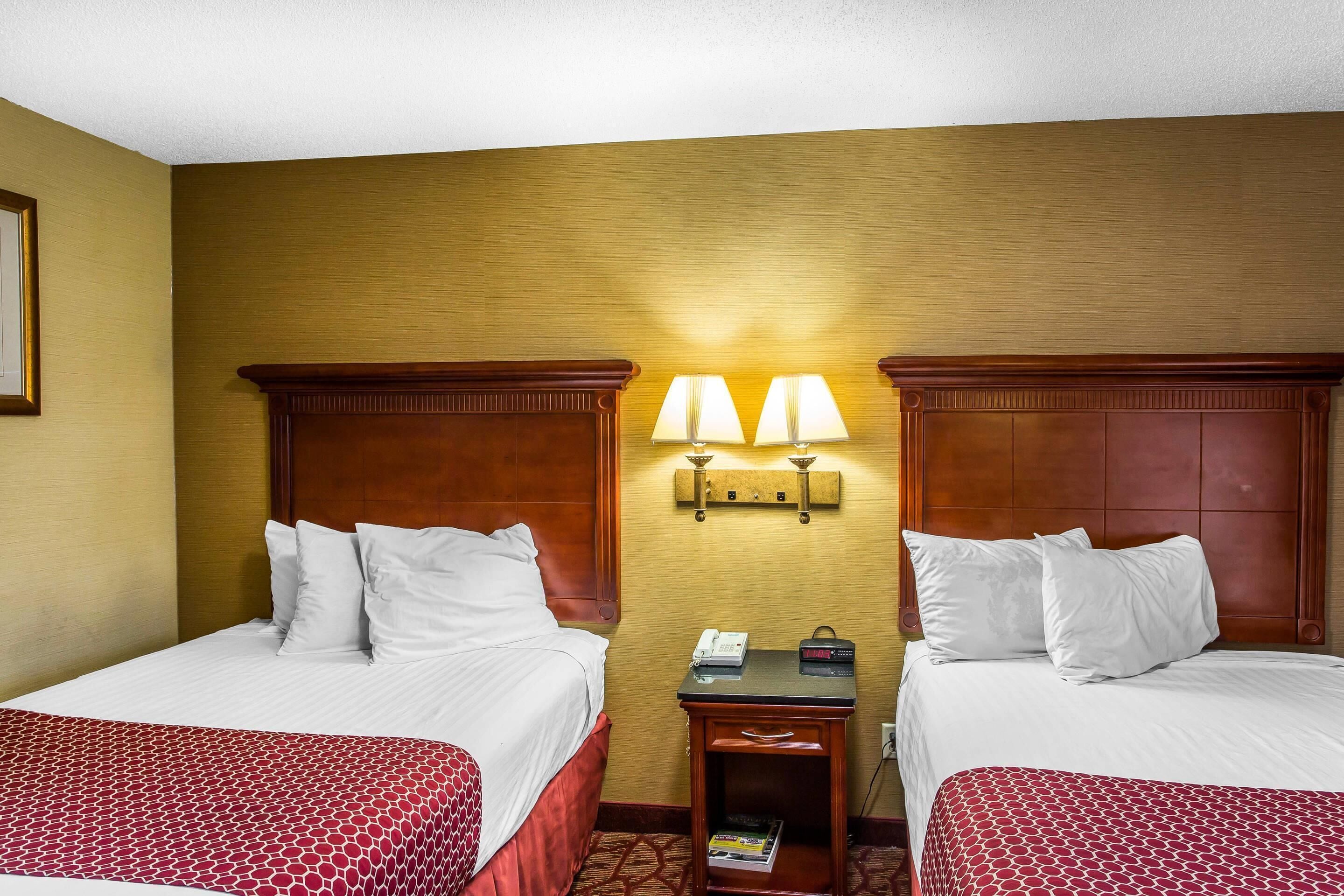 A hotel room with two beds, a nightstand with a lamp, and a wall-mounted light fixture.
