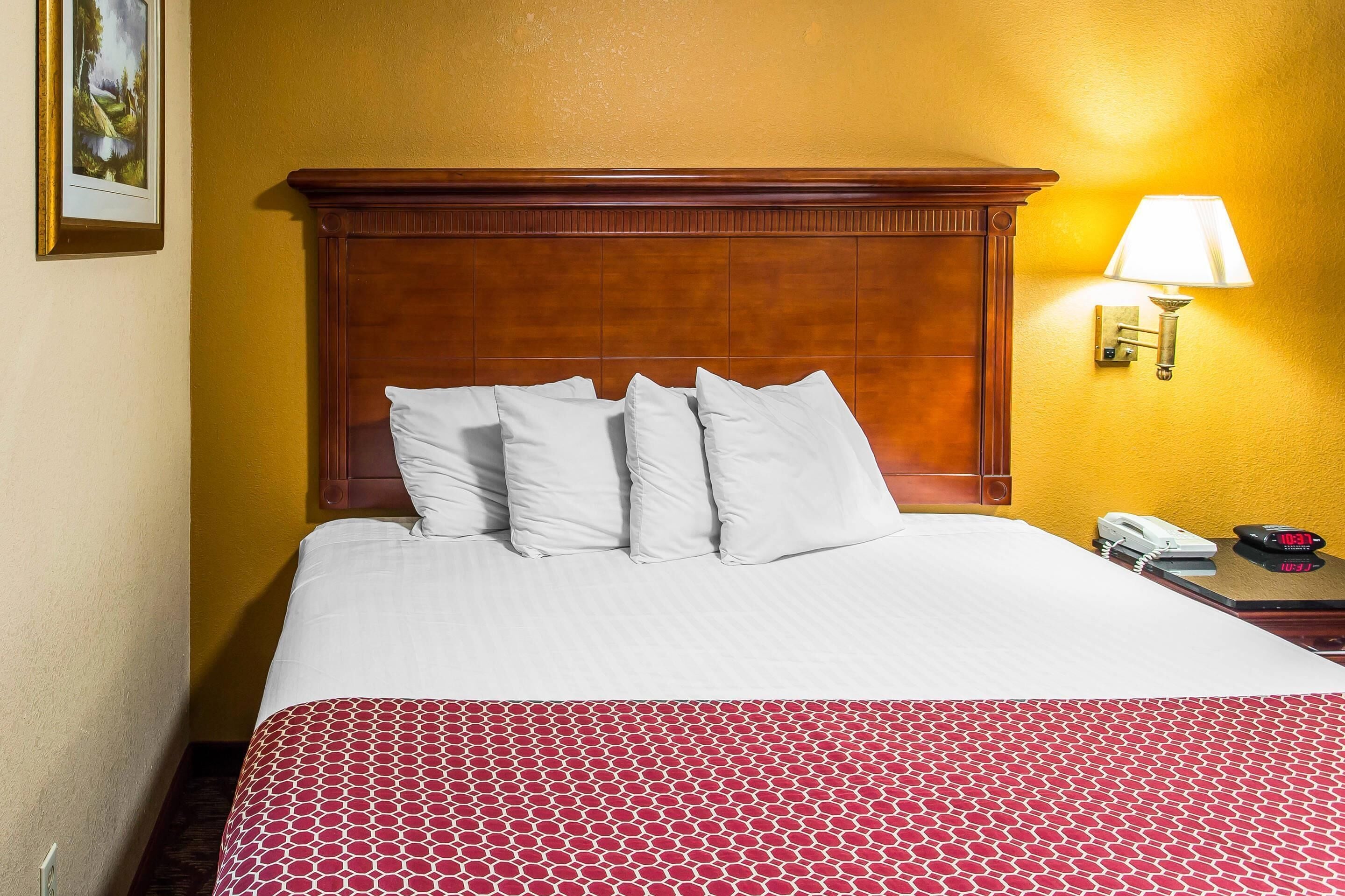 Habitación de hotel con cabecera de madera, cama con ropa blanca, colcha estampada roja, mesita de noche con lámpara y teléfono.