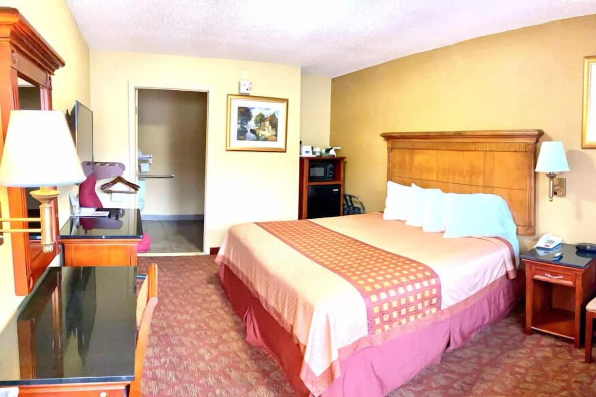 A hotel room with a large bed, a desk with a lamp, a wardrobe, a mirror, and a door leading to another room.