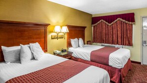 Ein Hotelzimmer mit zwei Betten, einem Holz-Kopfteil, einem Nachttisch mit Lampe, einem Fenster mit Vorhängen und einer Tür.