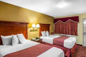 Una habitación de hotel con dos camas, cabecero de madera, mesita de noche con lámpara, ventana con cortinas y una puerta.