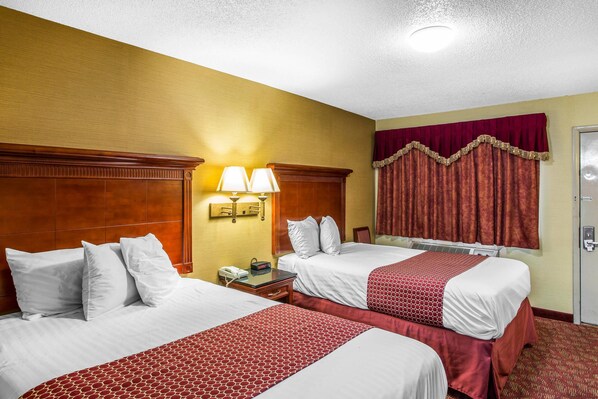 A hotel room with two beds, a wooden headboard, a nightstand with a lamp, a window with curtains, and a door.