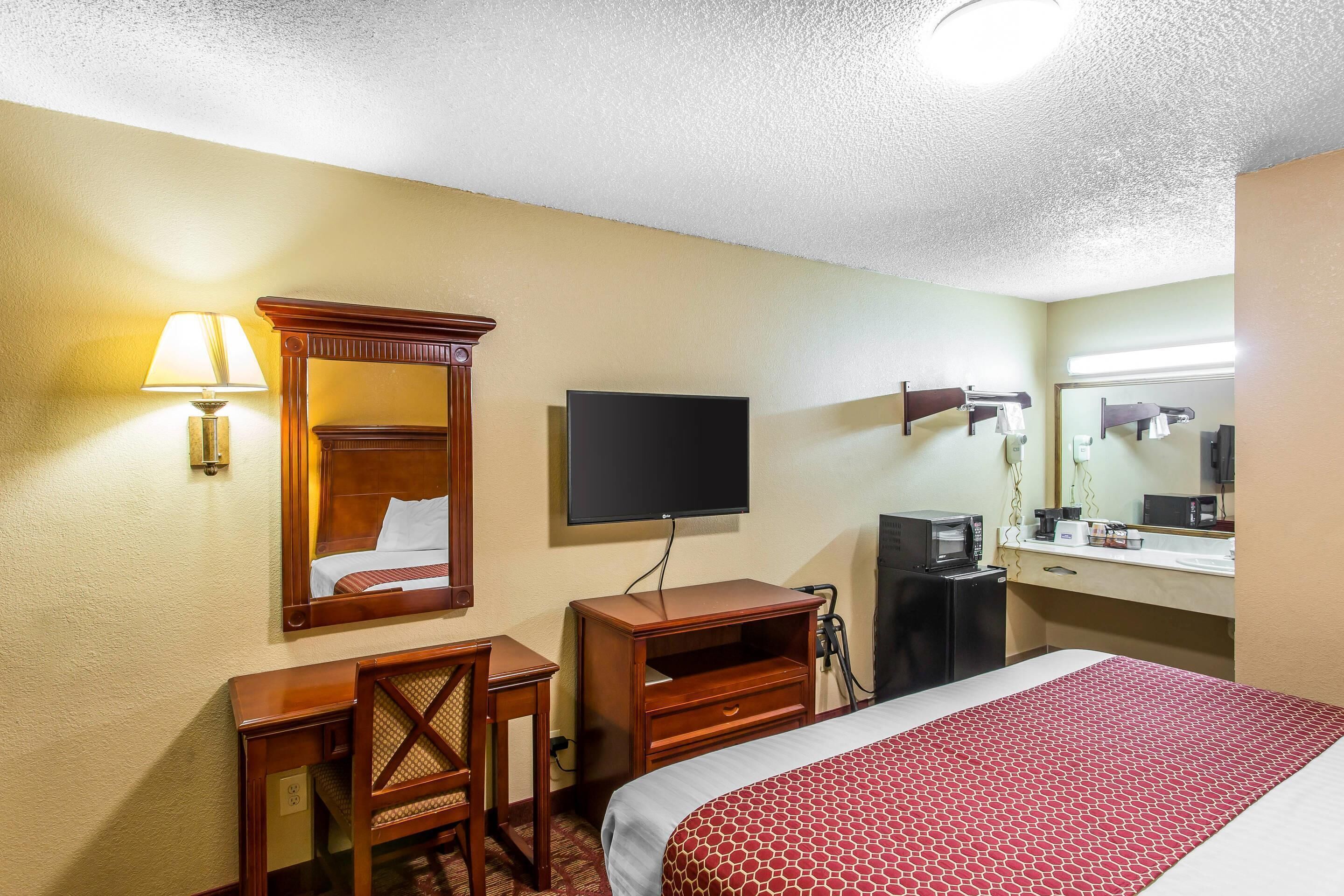 A hotel room with a bed, a desk, a television, a mirror, and a small kitchenette.