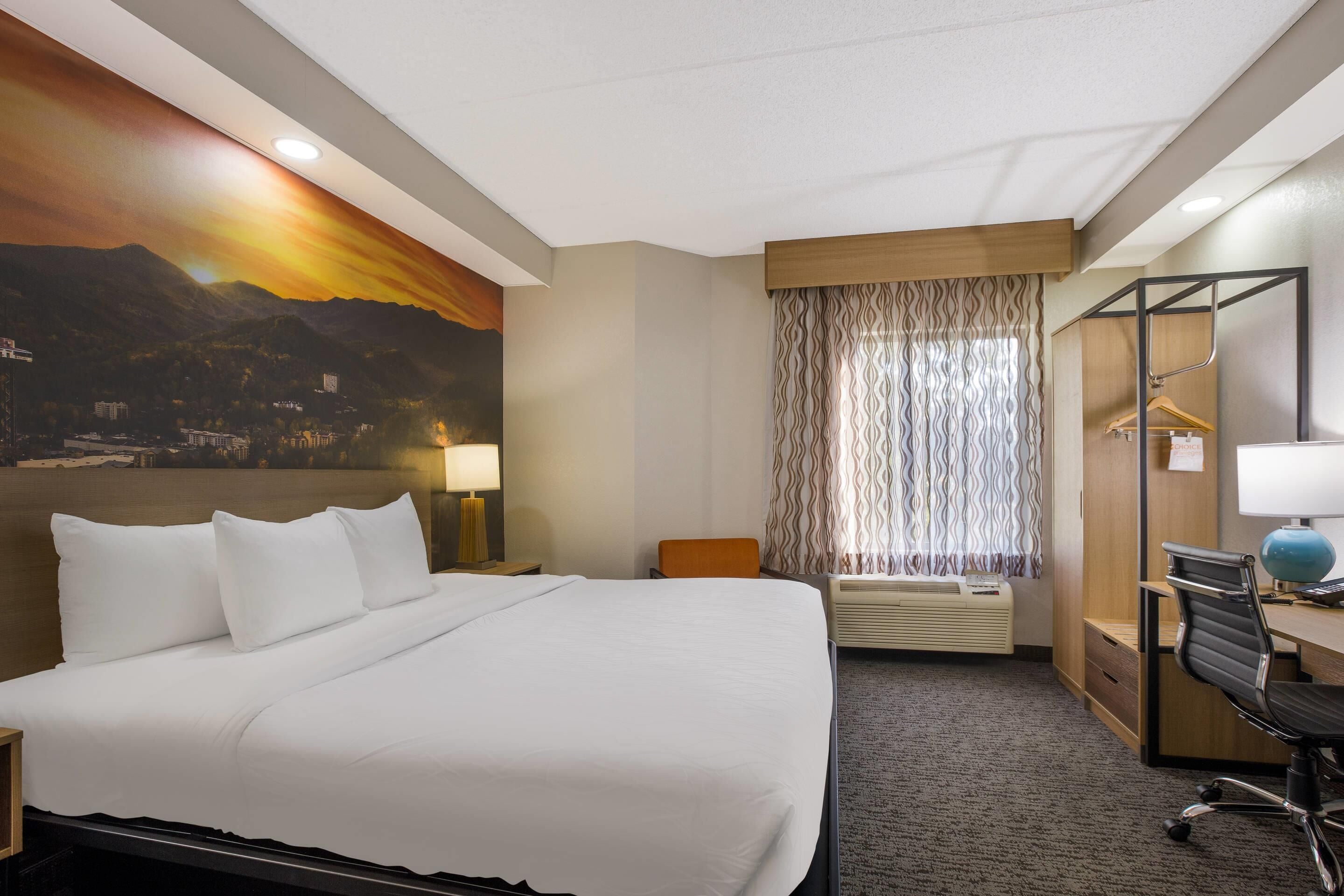 Ein Hotelzimmer mit einem großen Bett, einem Schreibtisch, einem Stuhl, einer Schreibtischlampe, einer Hängelampe, einem Fenster mit Vorhängen und einer Wand mit Stadtansicht-Mural.