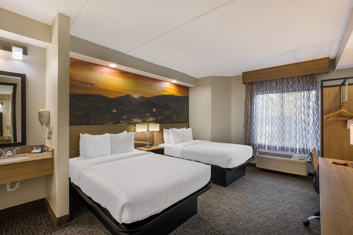 A hotel room with two beds, a large mirror, a sink, and a painting of a sunset.
