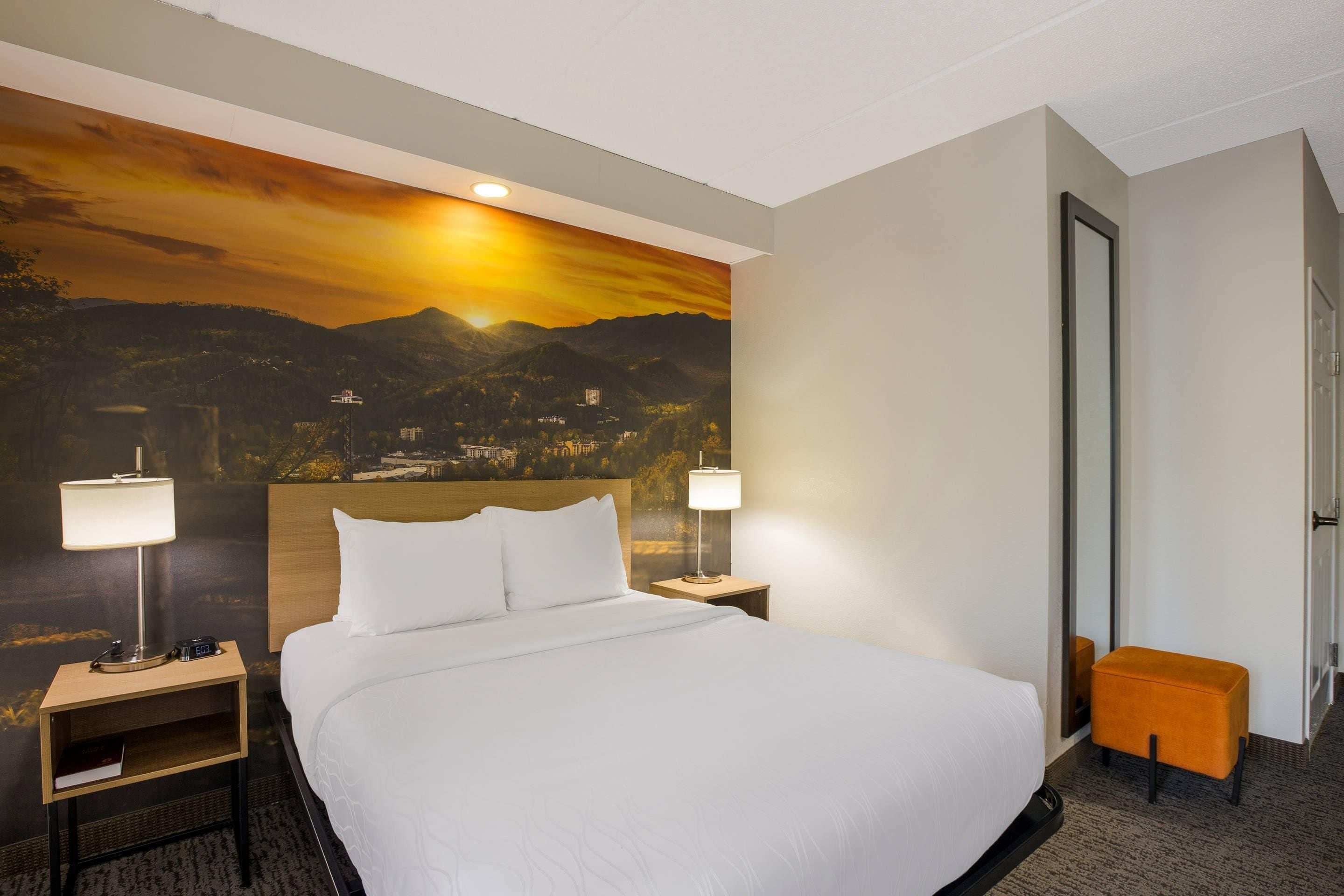 A hotel room with a large bed, two bedside tables, a nightstand, a small orange ottoman, and a wall mural of a sunset over a mountainous landscape.