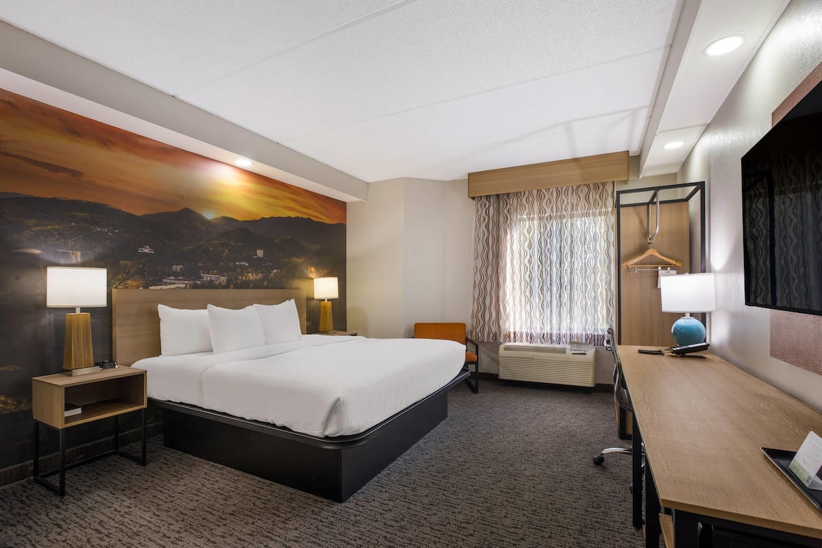 A hotel room with a large bed, two bedside tables with lamps, a desk with a chair, a window with curtains, and a wall mural.