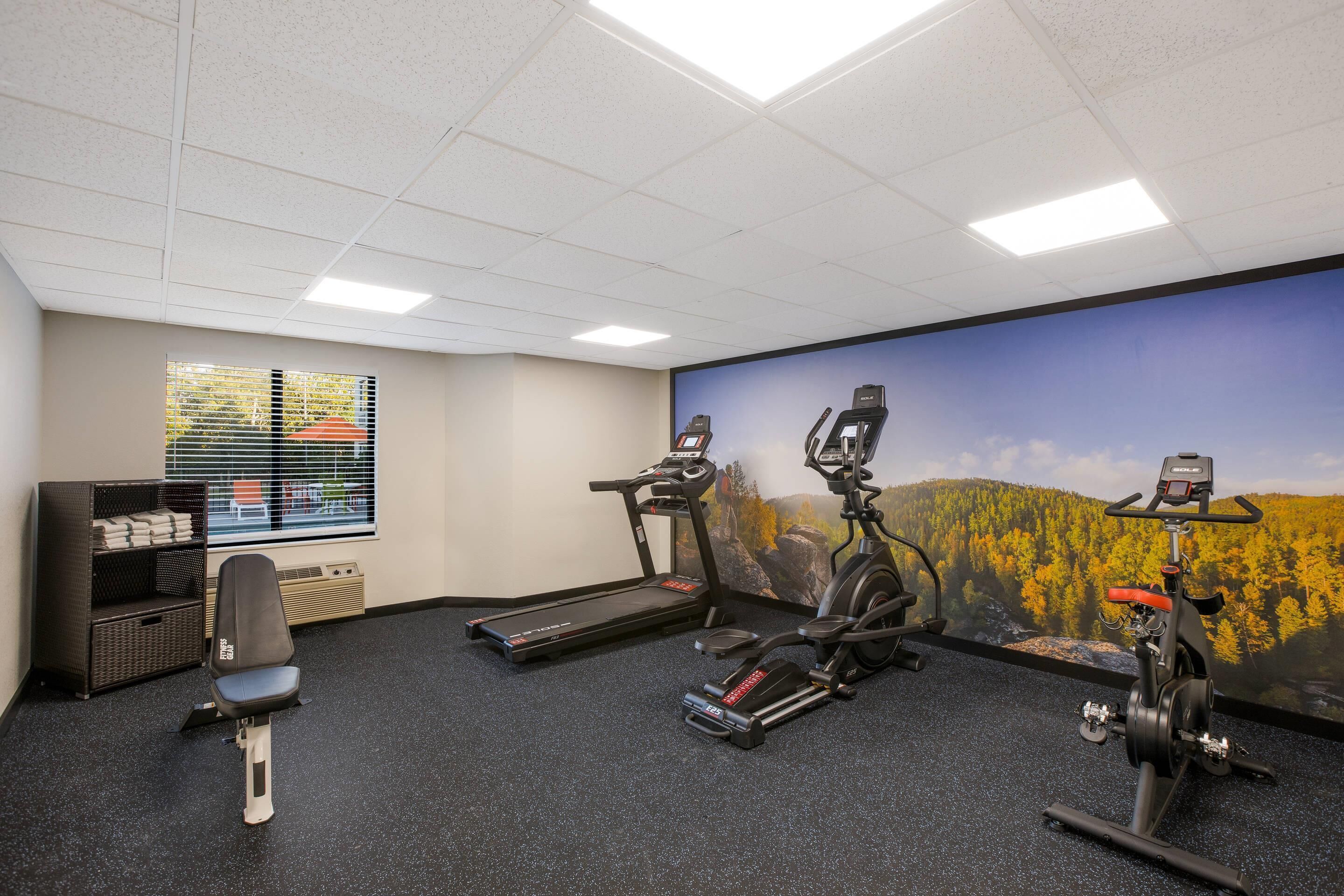 A fitness room with a treadmill, elliptical, and stationary bike, a wall-mounted TV, and a window with blinds.