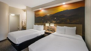 A hotel room with two beds, a nightstand with a lamp, a bathroom with a mirror and towel rack, and a large painting of a sunset over mountains. - Clarion Pointe Sevierville-Pigeon Forge (Sevierville)