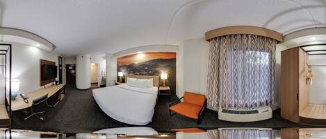 A panoramic view of a hotel room with a curved bed, a desk with a chair, a TV, and a large window with curtains.
