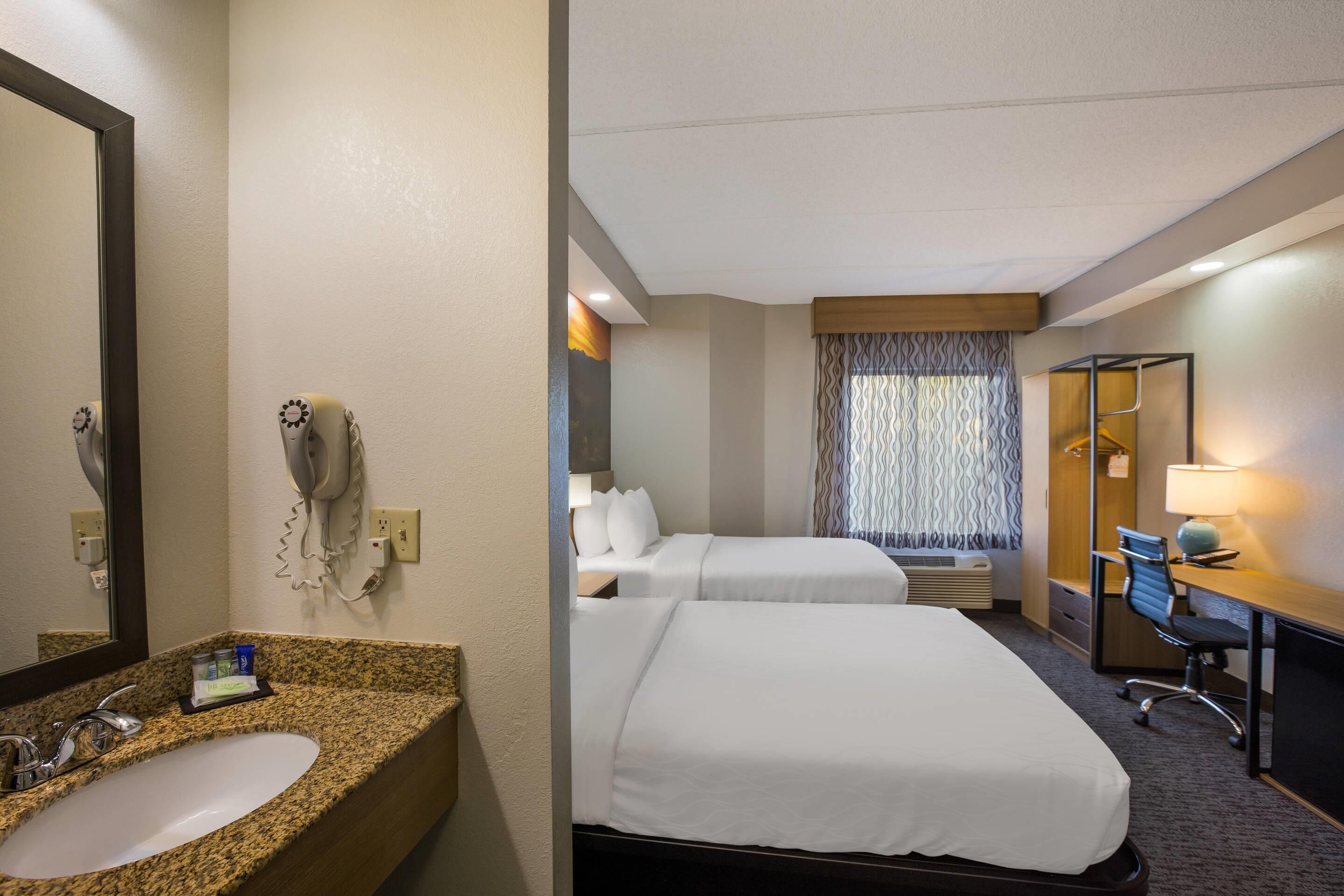 A hotel room with a granite countertop sink, a mirror, a hairdryer, two beds with white linens, a desk with a lamp, and a patterned curtain.