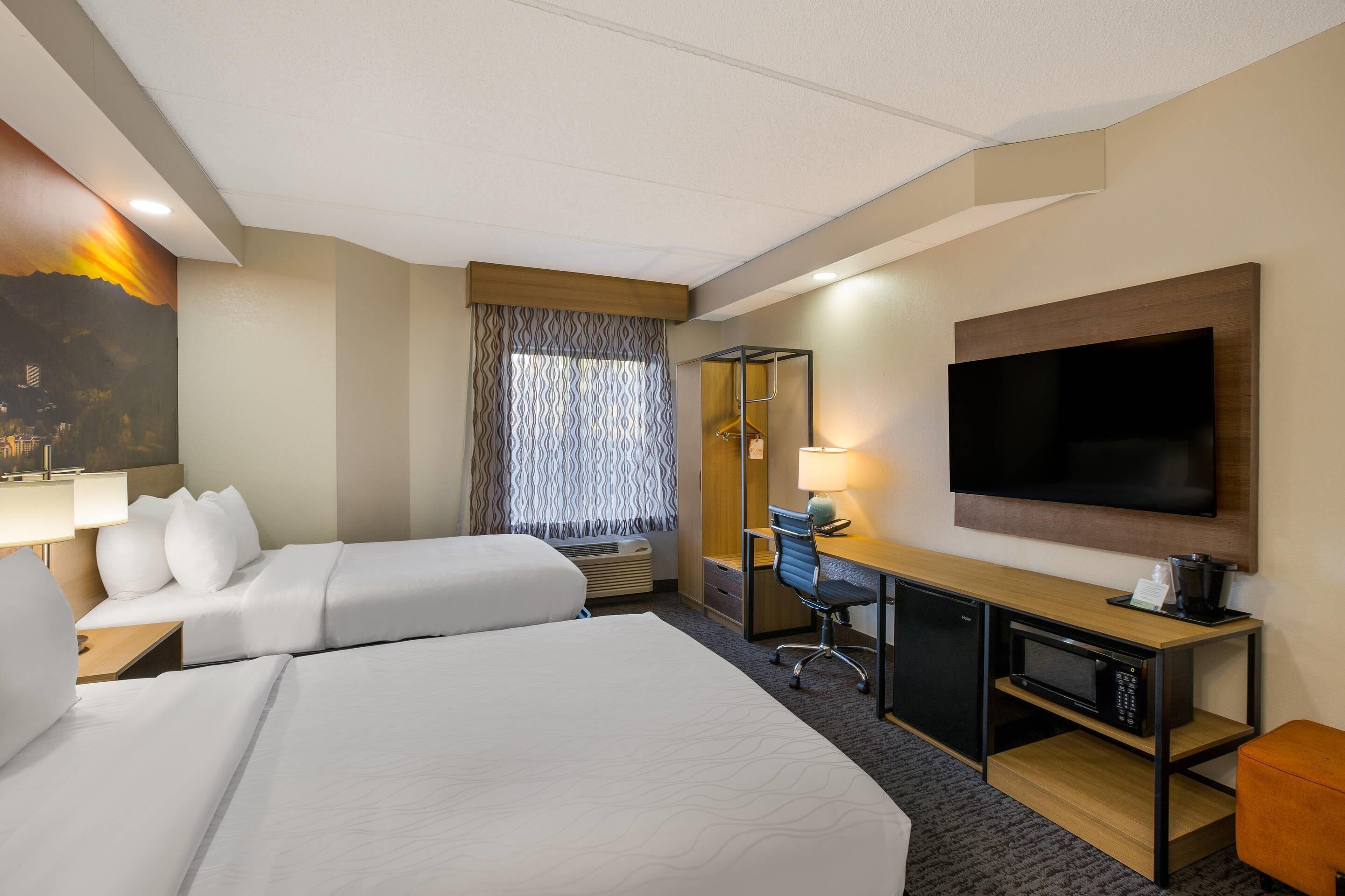 A hotel room with two beds, a desk, a TV, and a large window with patterned curtains.