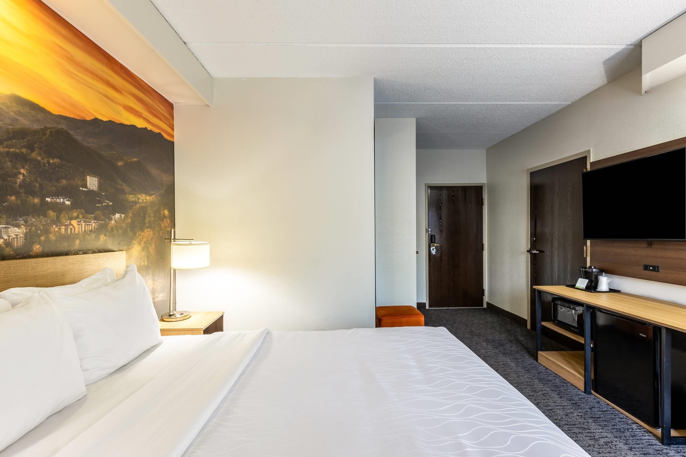 A hotel room with a bed, a desk, a TV, and a painting of a mountain landscape.