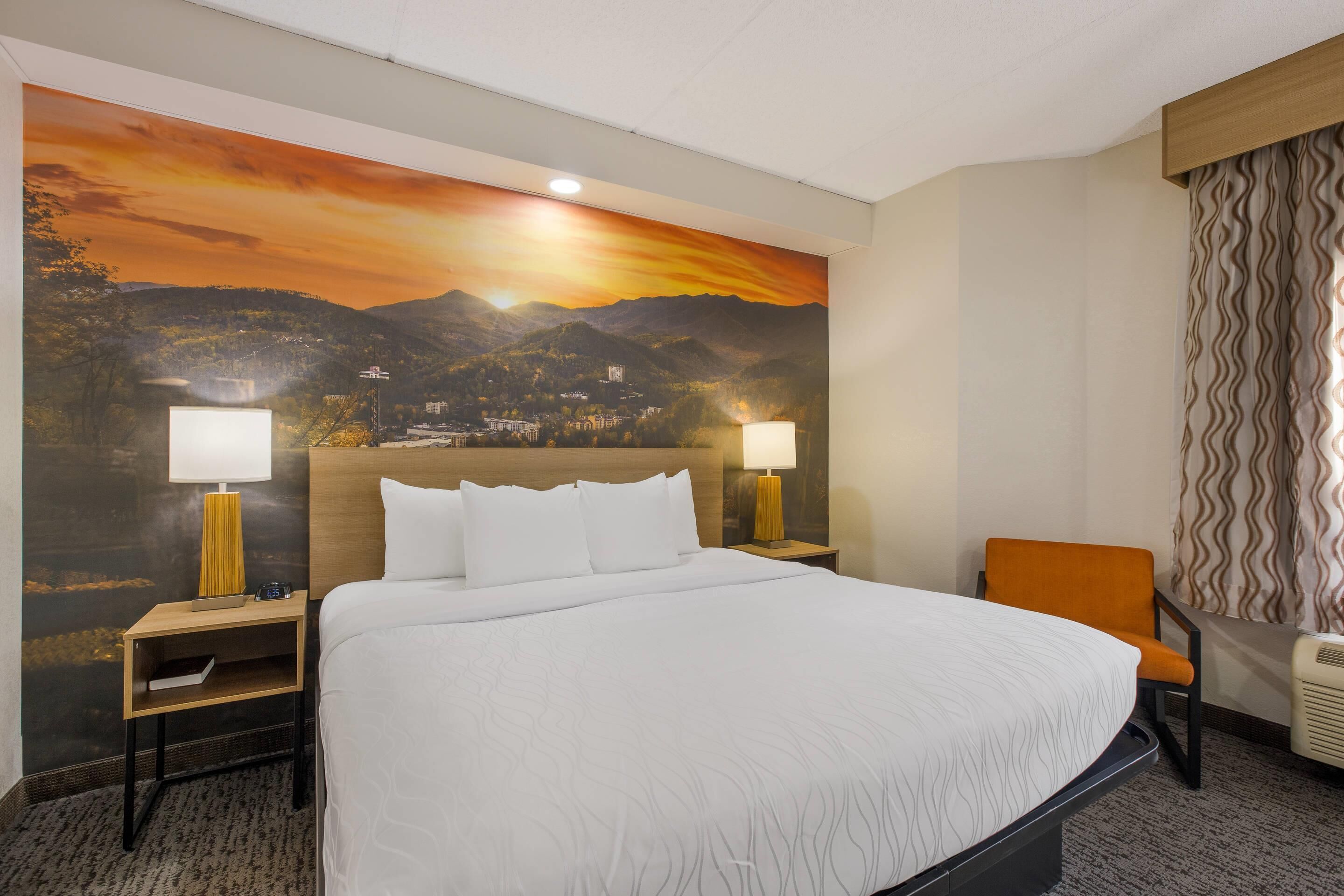 A hotel room with a large bed, two bedside lamps, a chair, a nightstand, and a wall mural of a sunset over mountains.