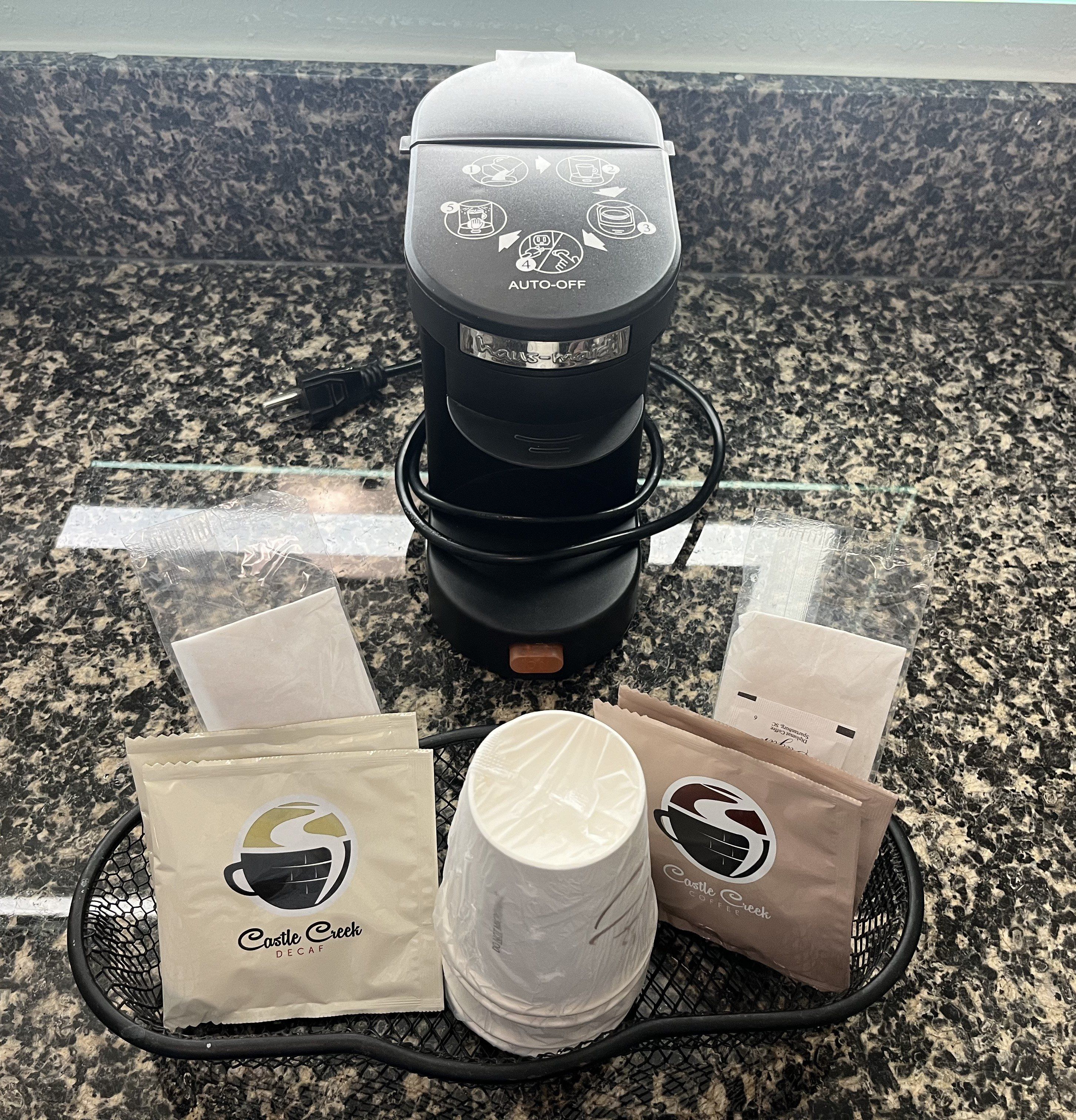 Uma cafeteira, uma xícara de café e dois sacos de café em uma bancada de granito.