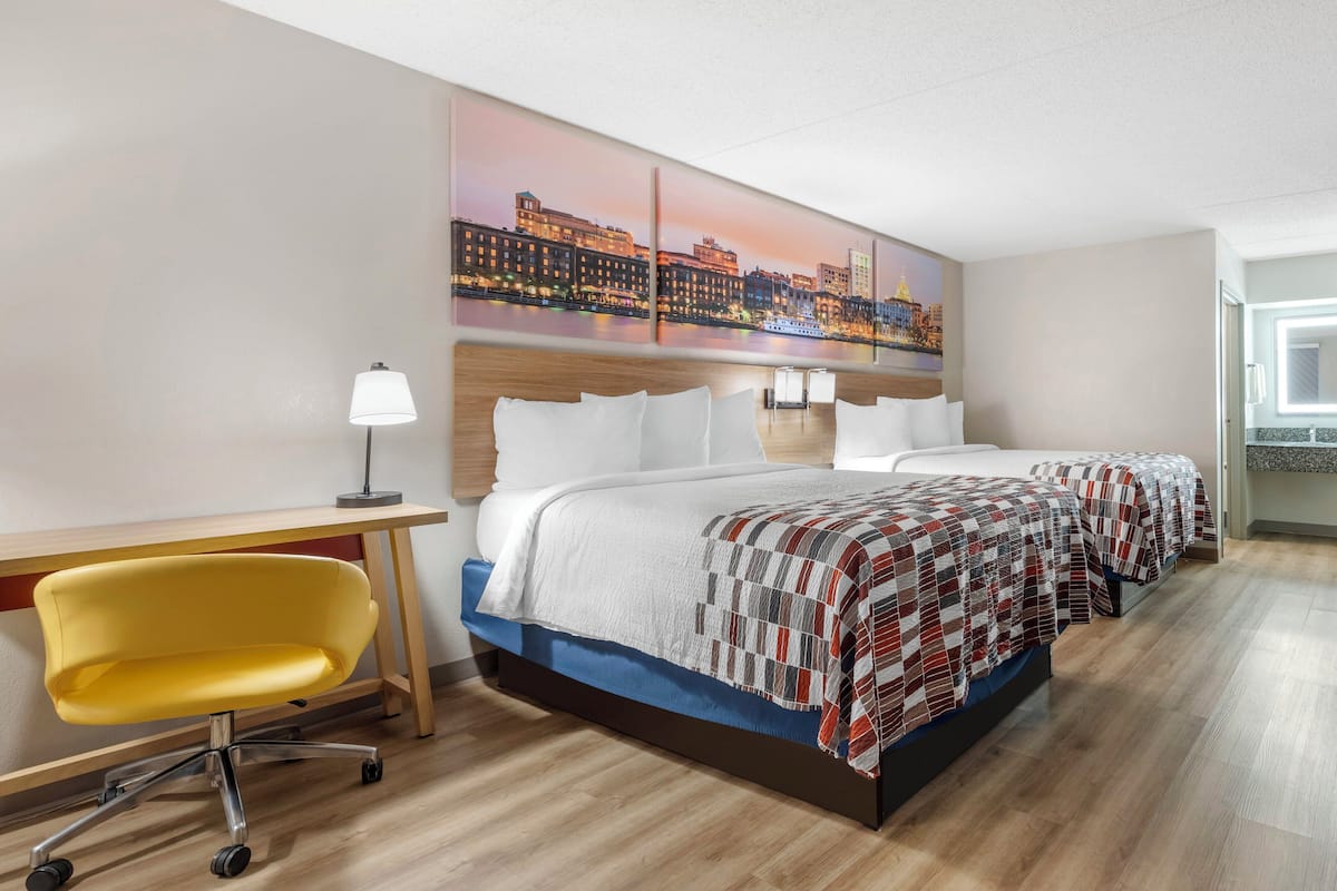A hotel room with a bed, a desk, a chair, and a cityscape painting on the wall.