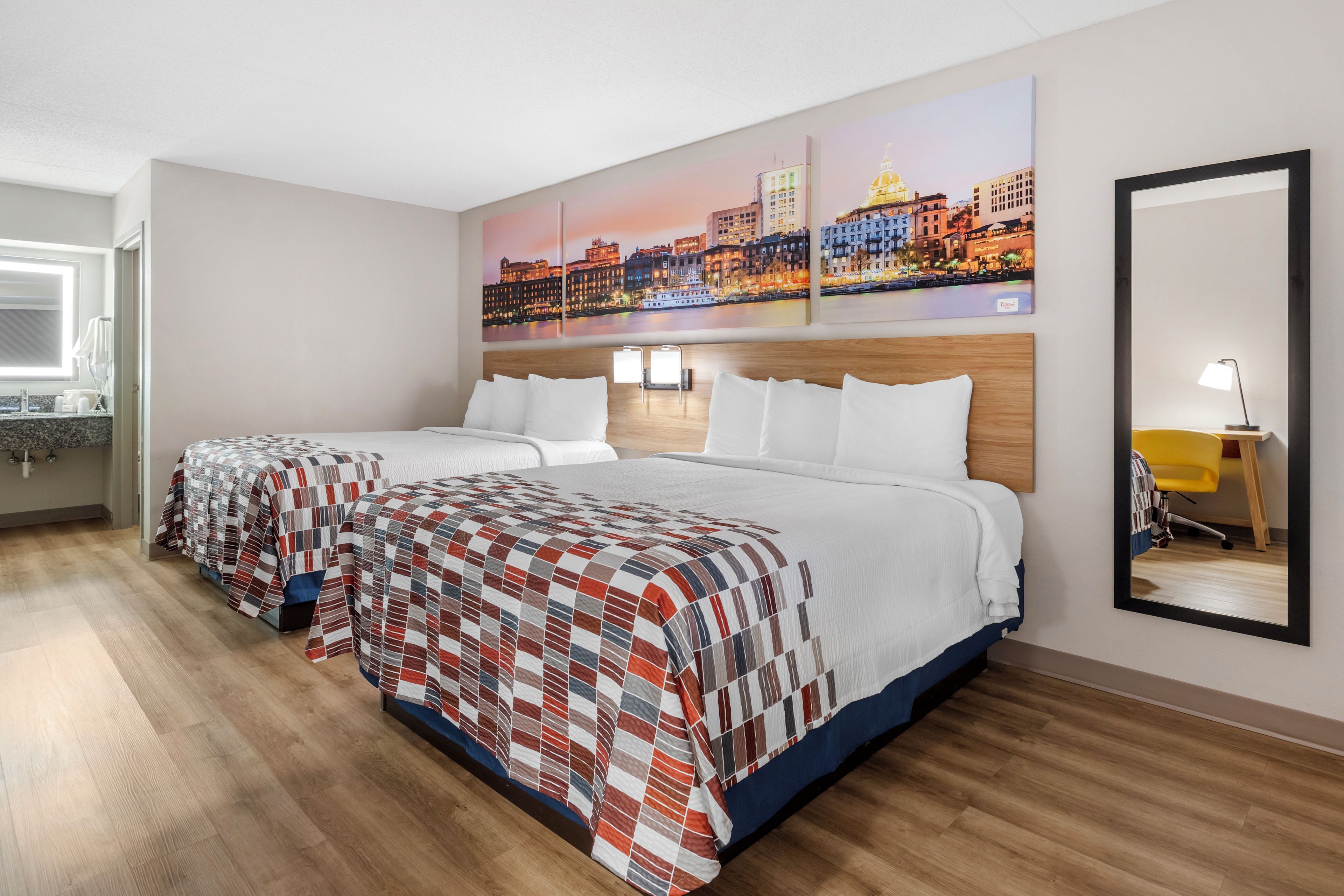 A hotel room with two beds, a cityscape painting, a mirror, and a desk with a chair.