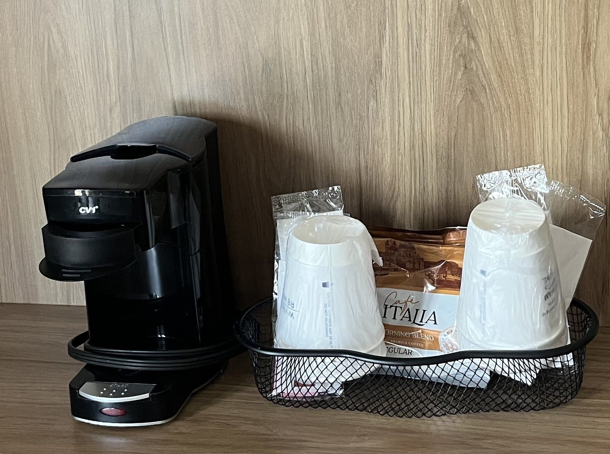 A black coffee maker on a table next to a basket containing two white coffee cups and a bag of coffee.