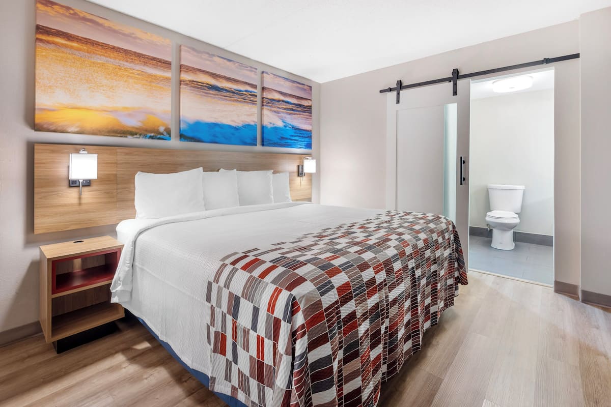A hotel room with a large bed, a wooden headboard, a nightstand with a lamp, a wall-mounted shelf, and a sliding door leading to a bathroom.
