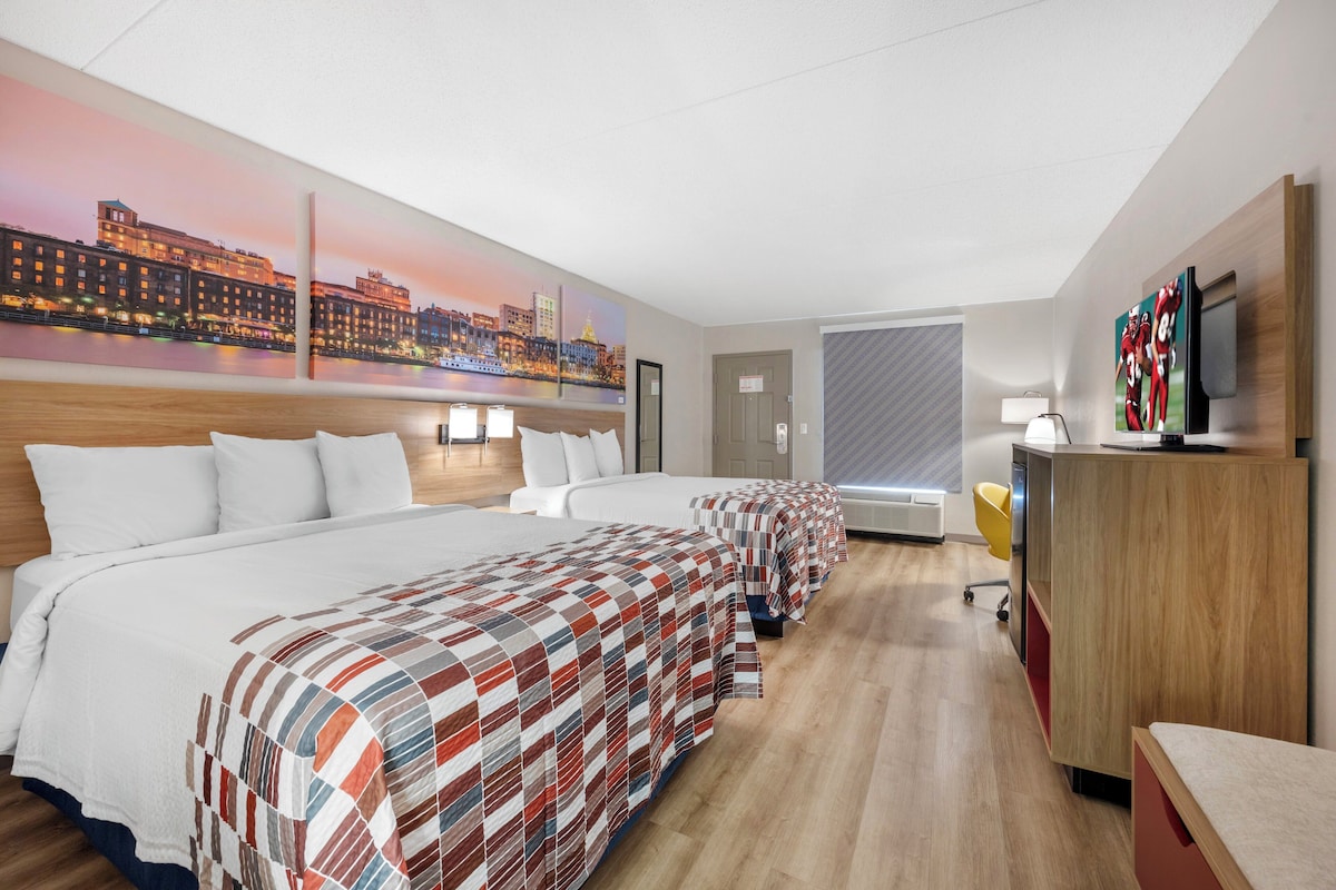Ein Hotelzimmer mit einem großen Bett, zwei Einzelbetten, einem Schreibtisch mit Fernseher und einem Stadtbild an der Wand.