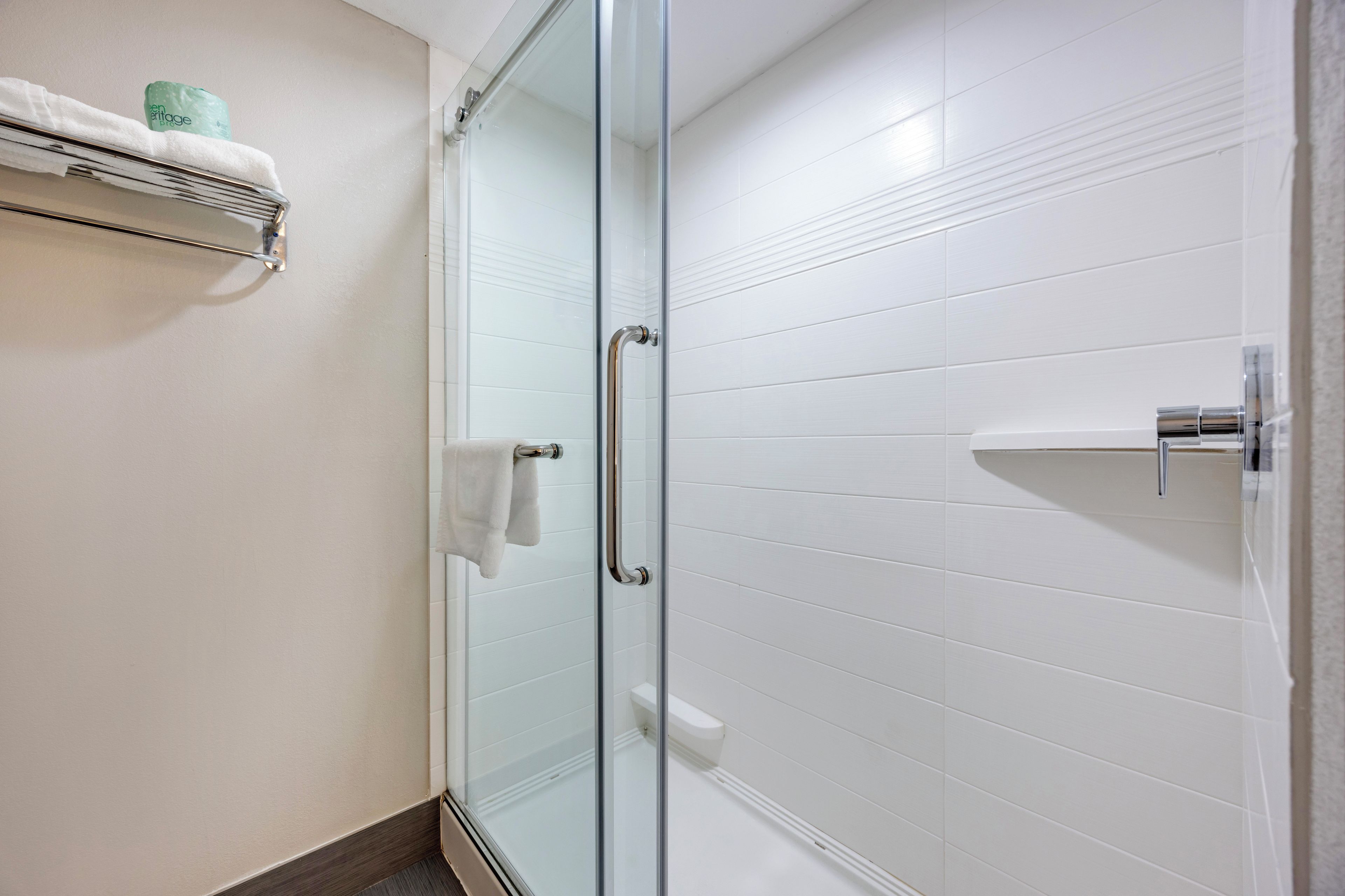 Une salle de bain avec une porte de douche en verre, des murs recouverts de carreaux blancs, un porte-serviettes et un support à papier toilette.