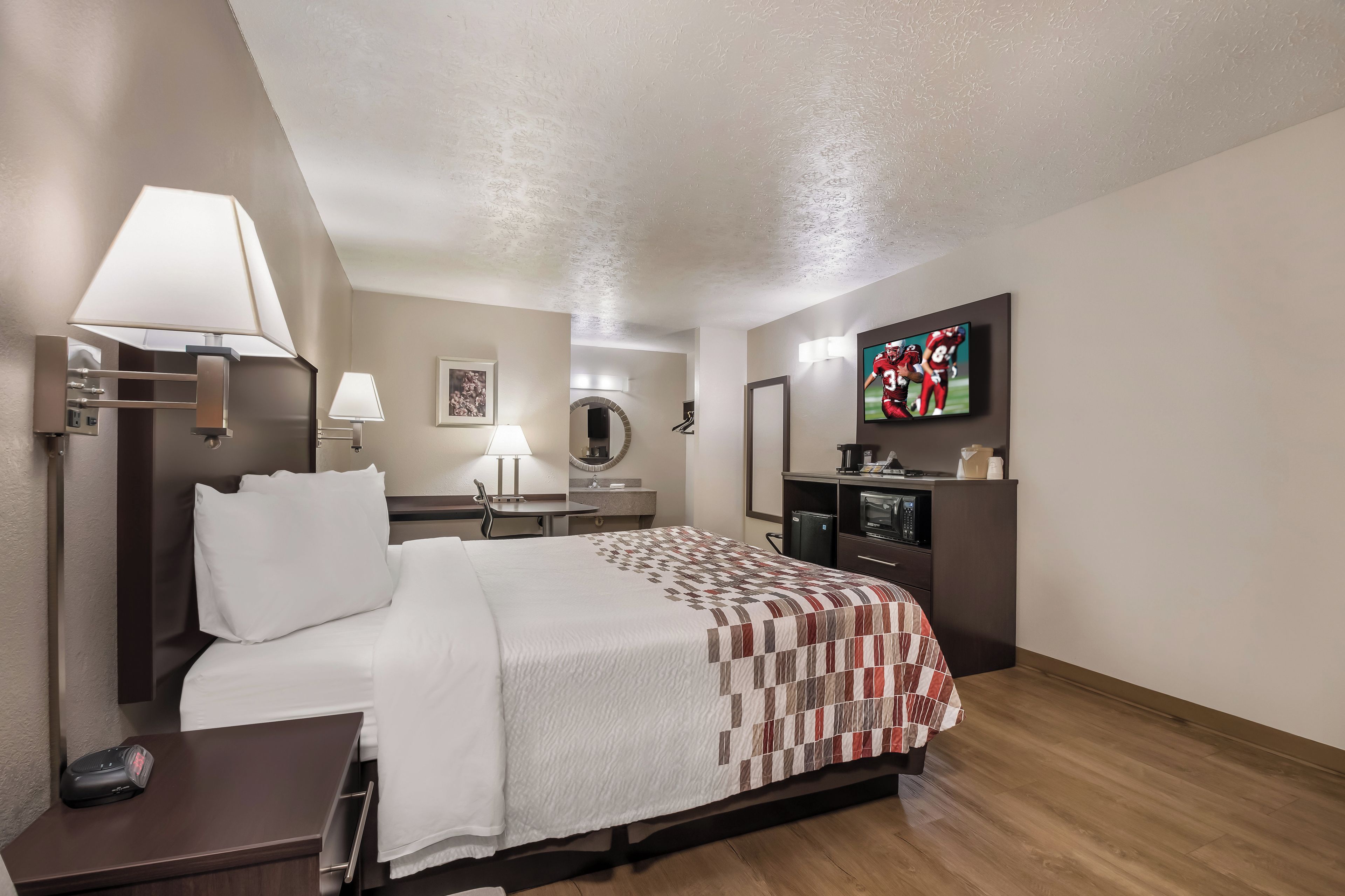 Ein Hotelzimmer mit Bett, Nachttisch, Lampe, Fernseher und einer kleinen Küchenzeile.