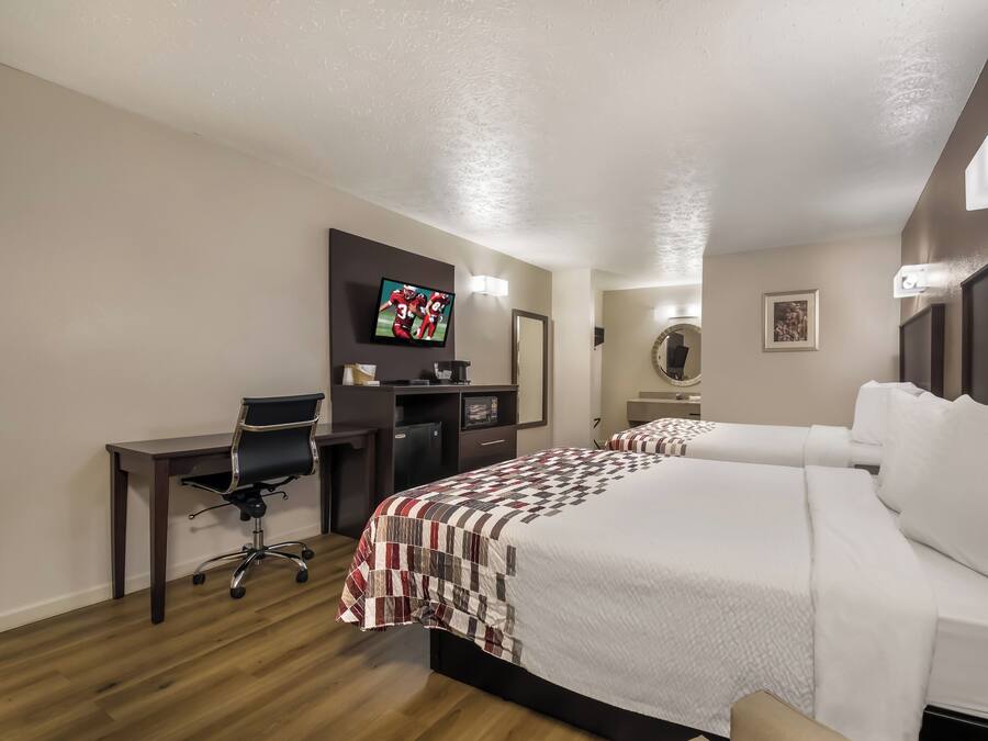 Quarto de hotel com cama, escrivaninha, cadeira, televisão e um pequeno frigorífico.
