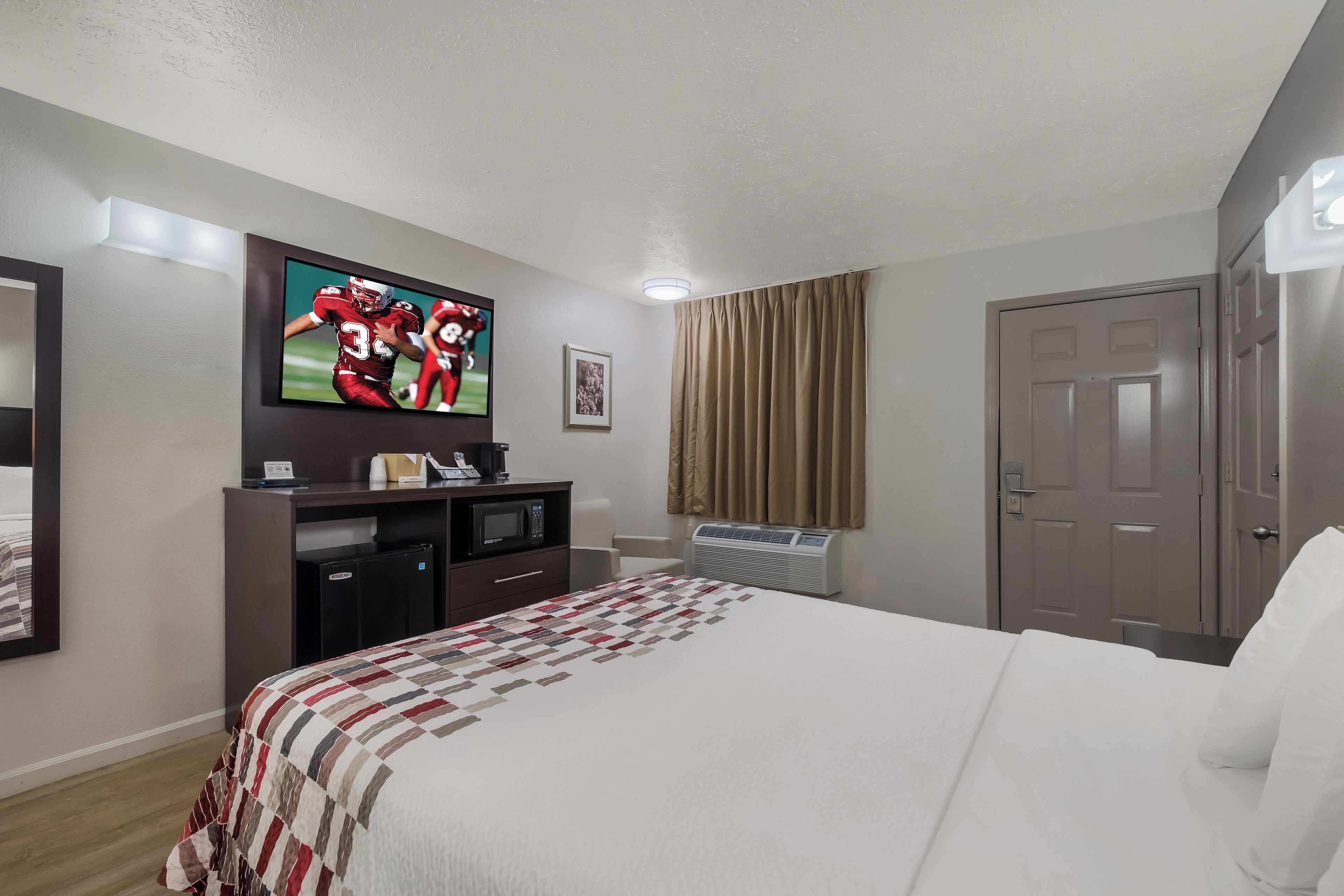 A hotel room with a bed, a flat-screen TV mounted on the wall, a desk with a microwave, and a door.