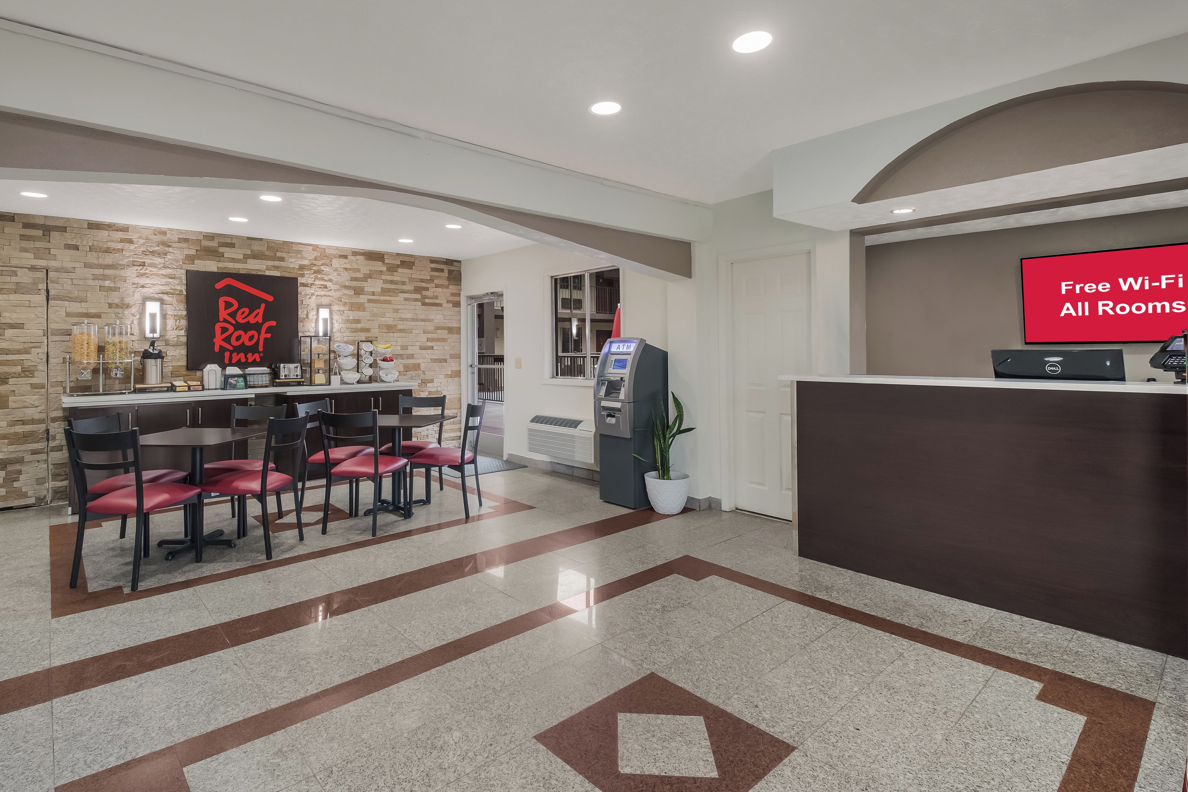 Un vestíbulo de hotel con mostrador de recepción, un letrero de Red Roof Inn, un cajero automático y un comedor con sillas y mesas.