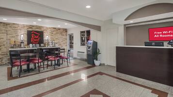 Un vestíbulo de hotel con mostrador de recepción, un letrero de Red Roof Inn, un cajero automático y un comedor con sillas y mesas.