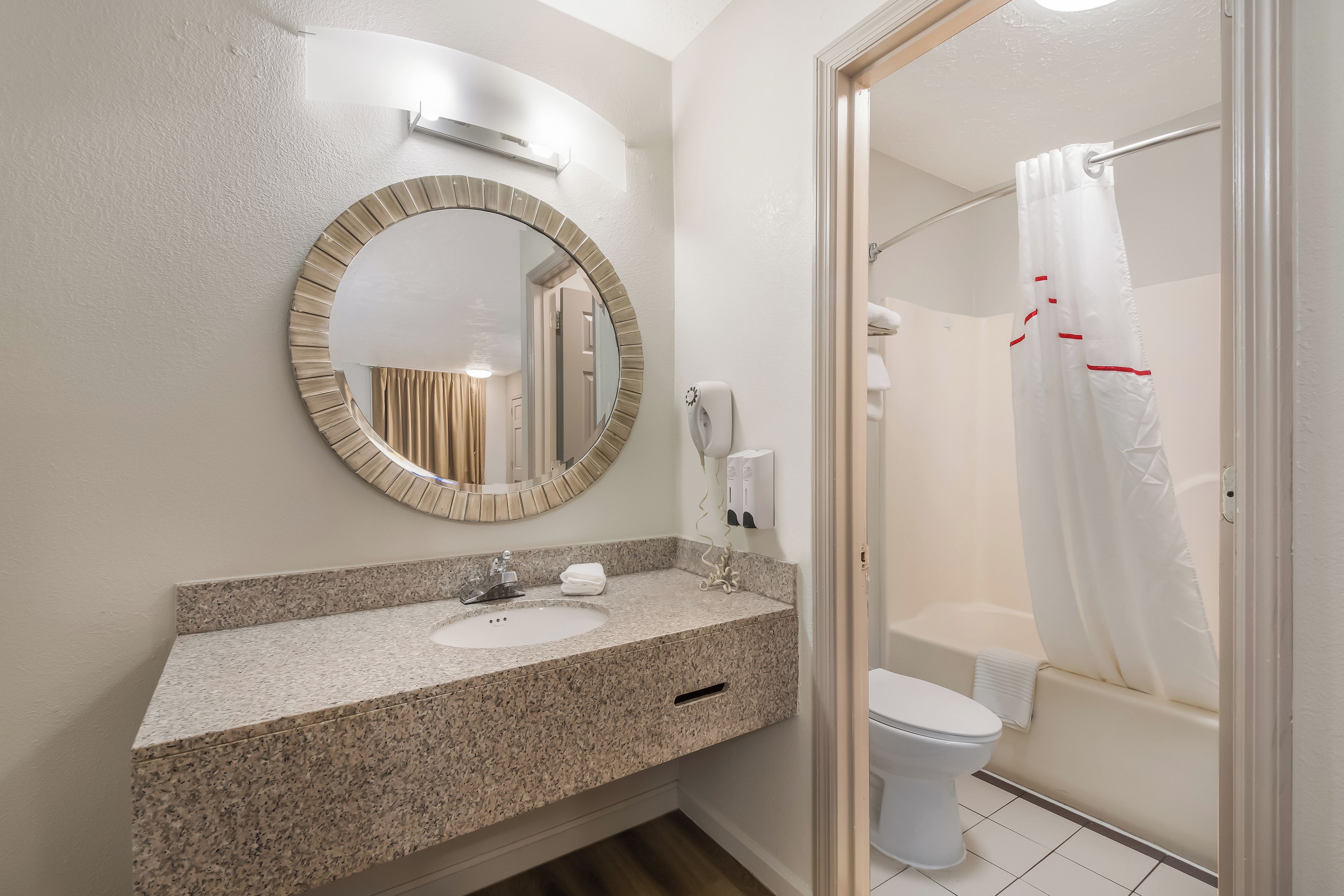 A bathroom with a granite countertop, a round mirror, a toilet, and a shower with a white curtain.