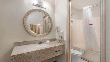 A bathroom with a granite countertop, a round mirror, a toilet, and a shower with a white curtain.