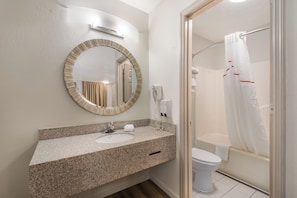 A bathroom with a granite countertop, a round mirror, a toilet, and a shower with a white curtain. - Red Roof Inn Crossville (Crossville)