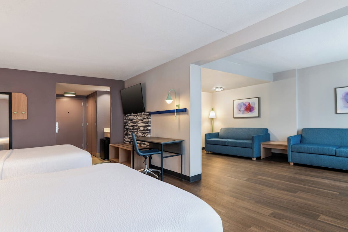 A hotel room with two beds, a desk with a chair, a TV, a blue sofa, and a painting on the wall.