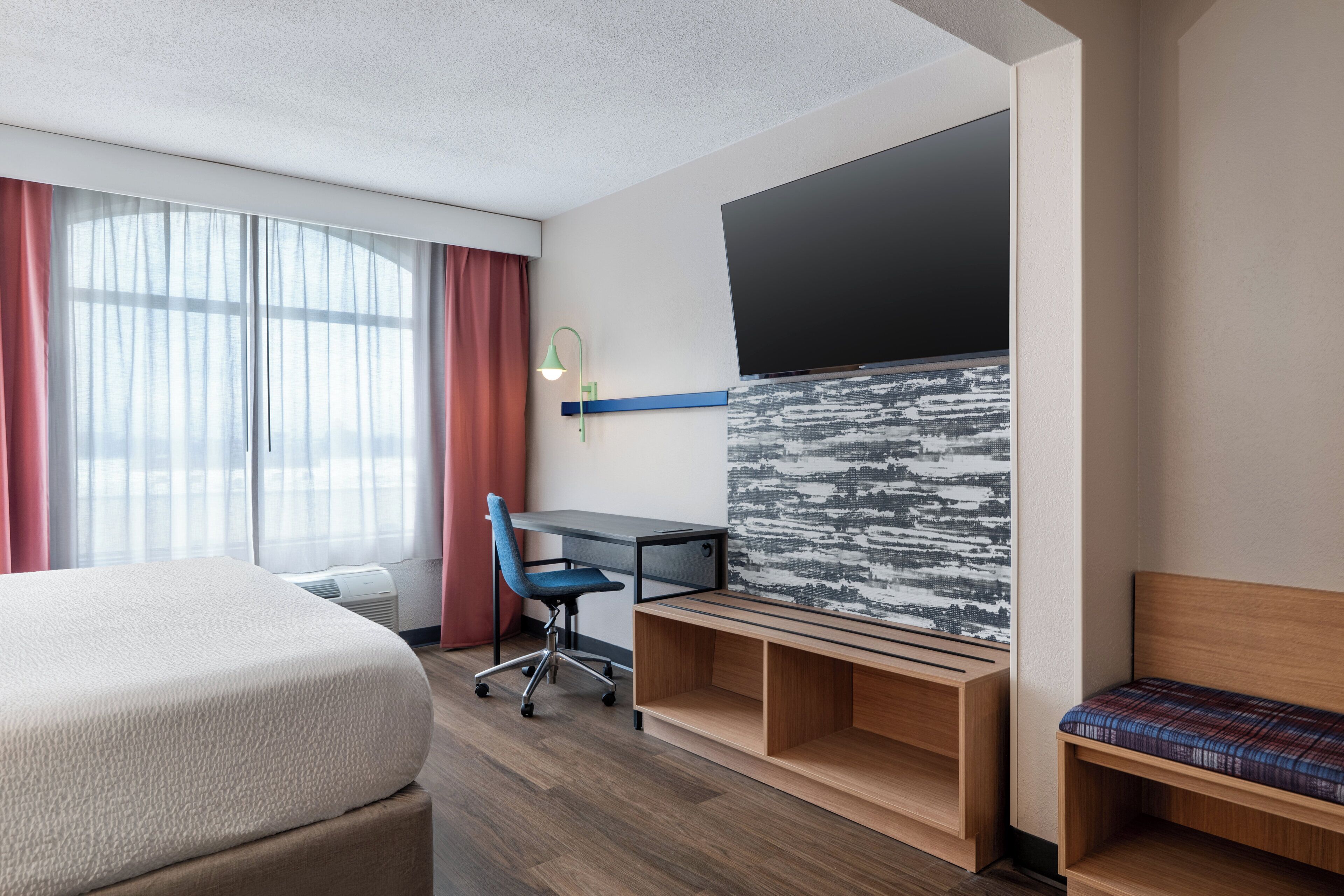 Habitación de hotel con cama, escritorio, silla, televisor y vistas al exterior.
