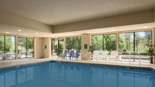 An indoor swimming pool with clear views of trees through large windows, surrounded by white lounge chairs and a few tables.