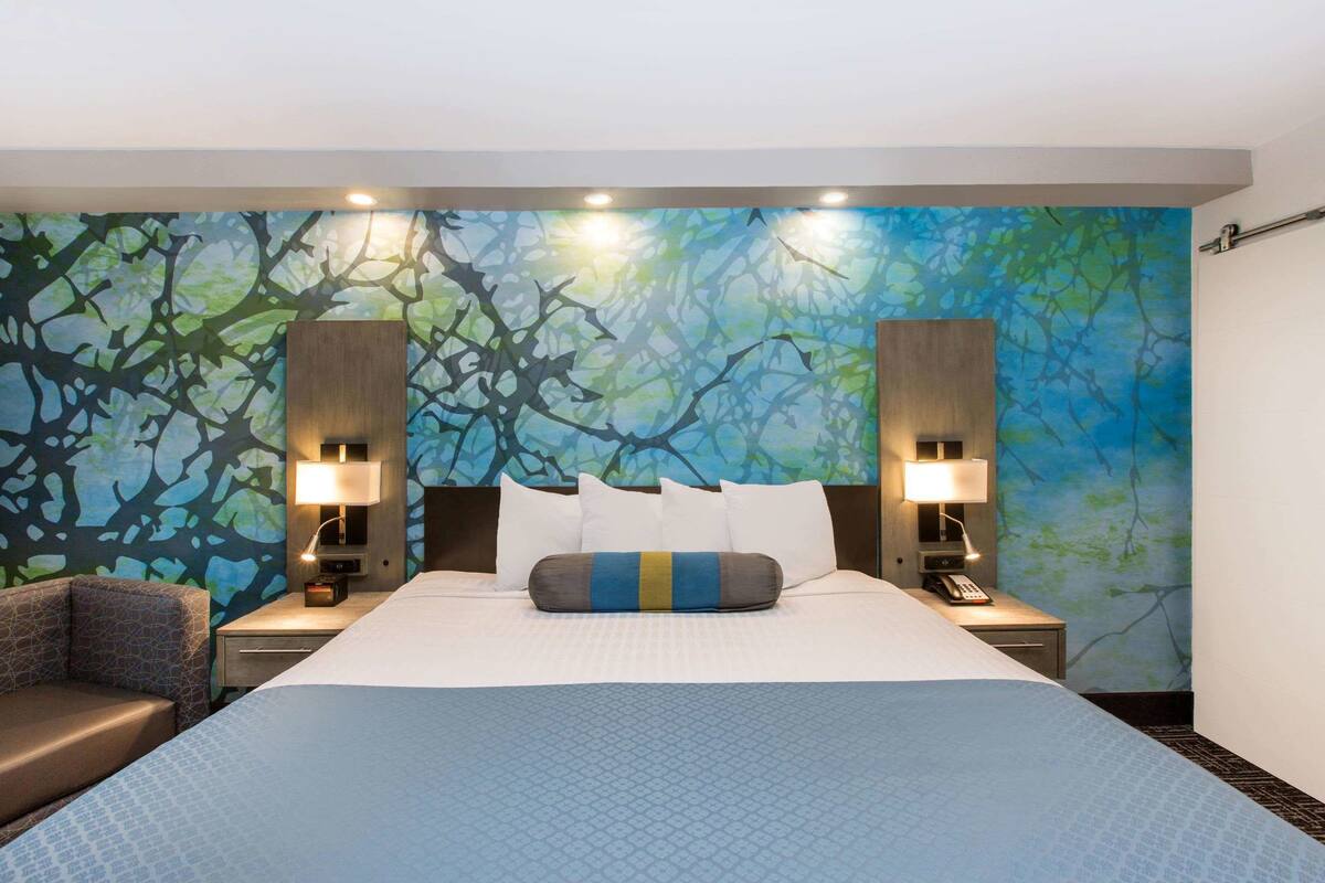 Habitación de hotel con una cama grande, dos mesitas de noche con lámparas, una silla y una pared decorada con un diseño de ramas de árbol.