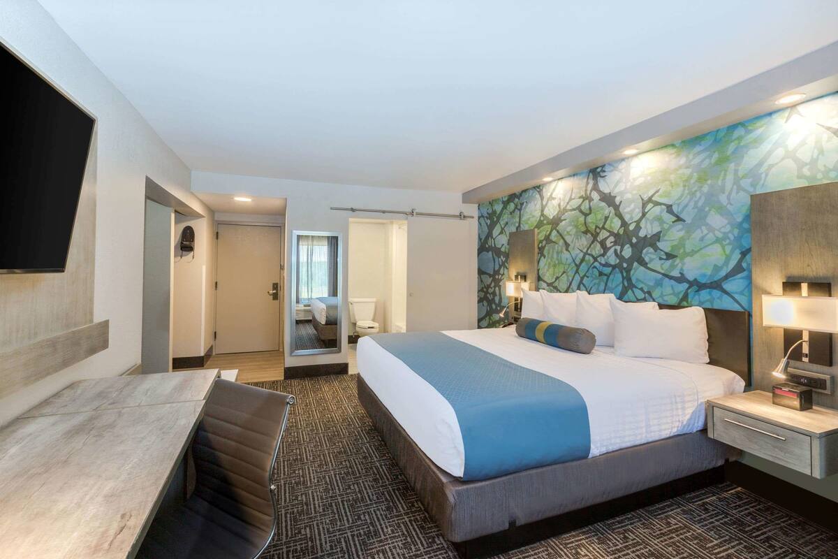 Habitación de hotel con una cama grande, un escritorio y un espejo. Hay un cuadro en la pared y un televisor fijado allí.