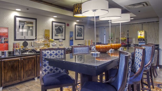 A hotel dining area with a large round table, blue patterned chairs, and a buffet with various food items. - Hampton Inn Bristol (Bristol)