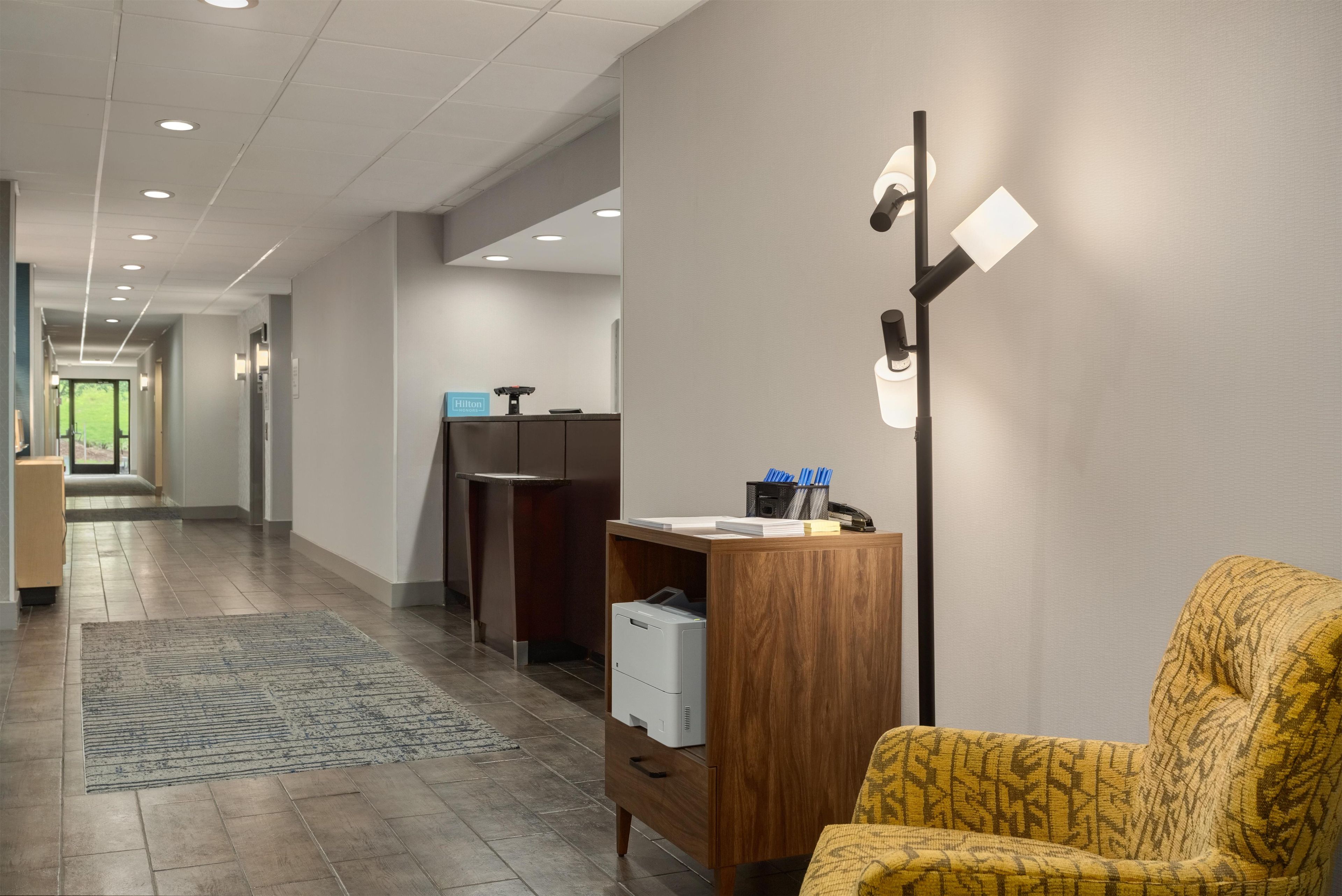Un hall d’hôtel avec un comptoir d’accueil, un bureau en bois avec une imprimante, un fauteuil jaune et une lampe sur pied.