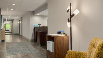 Un hall d’hôtel avec un comptoir d’accueil, un bureau en bois avec une imprimante, un fauteuil jaune et une lampe sur pied.