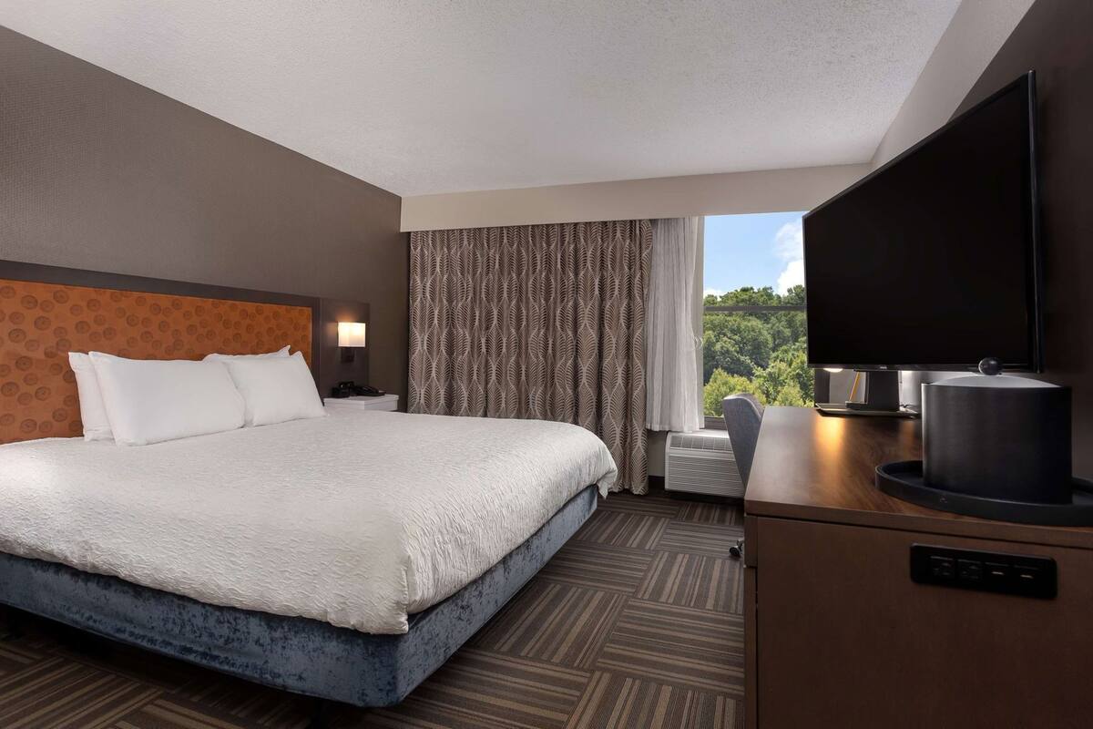 A hotel room with a bed, a desk with a flat-screen TV, a chair, and a window with a view.