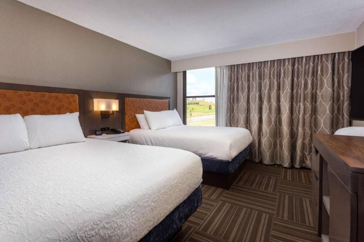 Habitación de hotel con dos camas, una mesita de noche, una lámpara, un escritorio y una ventana con cortinas.