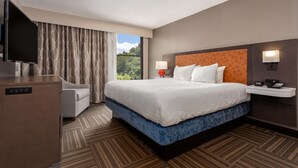 A hotel room with a large bed, a desk with a flat-screen TV, a sofa, a nightstand with a lamp, and a view of greenery outside. - Hampton Inn Bristol (Bristol)