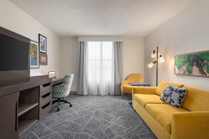 A hotel room with a desk, a yellow sofa, a chair, a TV, a painting, and a window with curtains.
