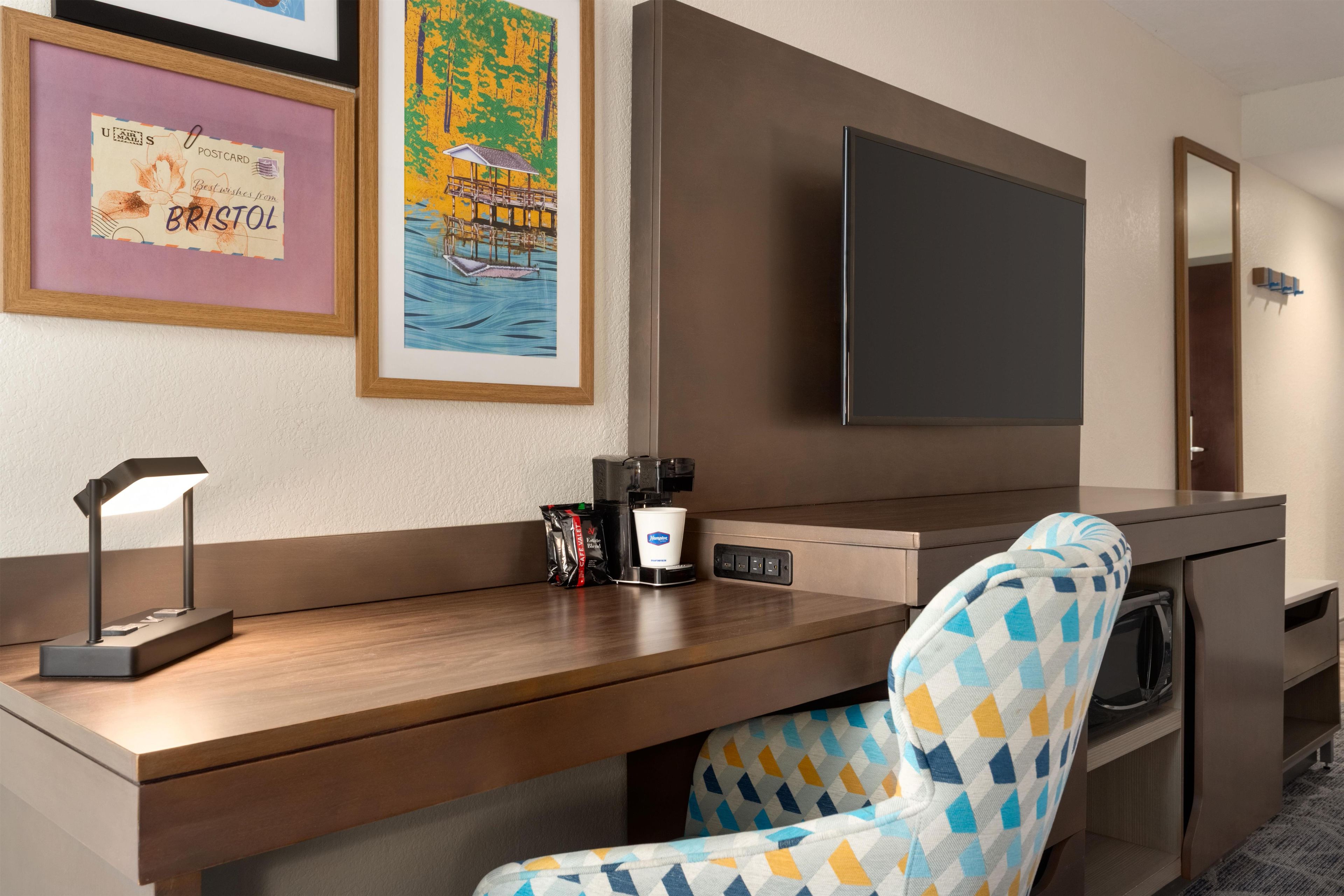 Habitación de hotel con un escritorio de madera, un televisor de pantalla plana montado en la pared, un sillón estampado y una cafetera.