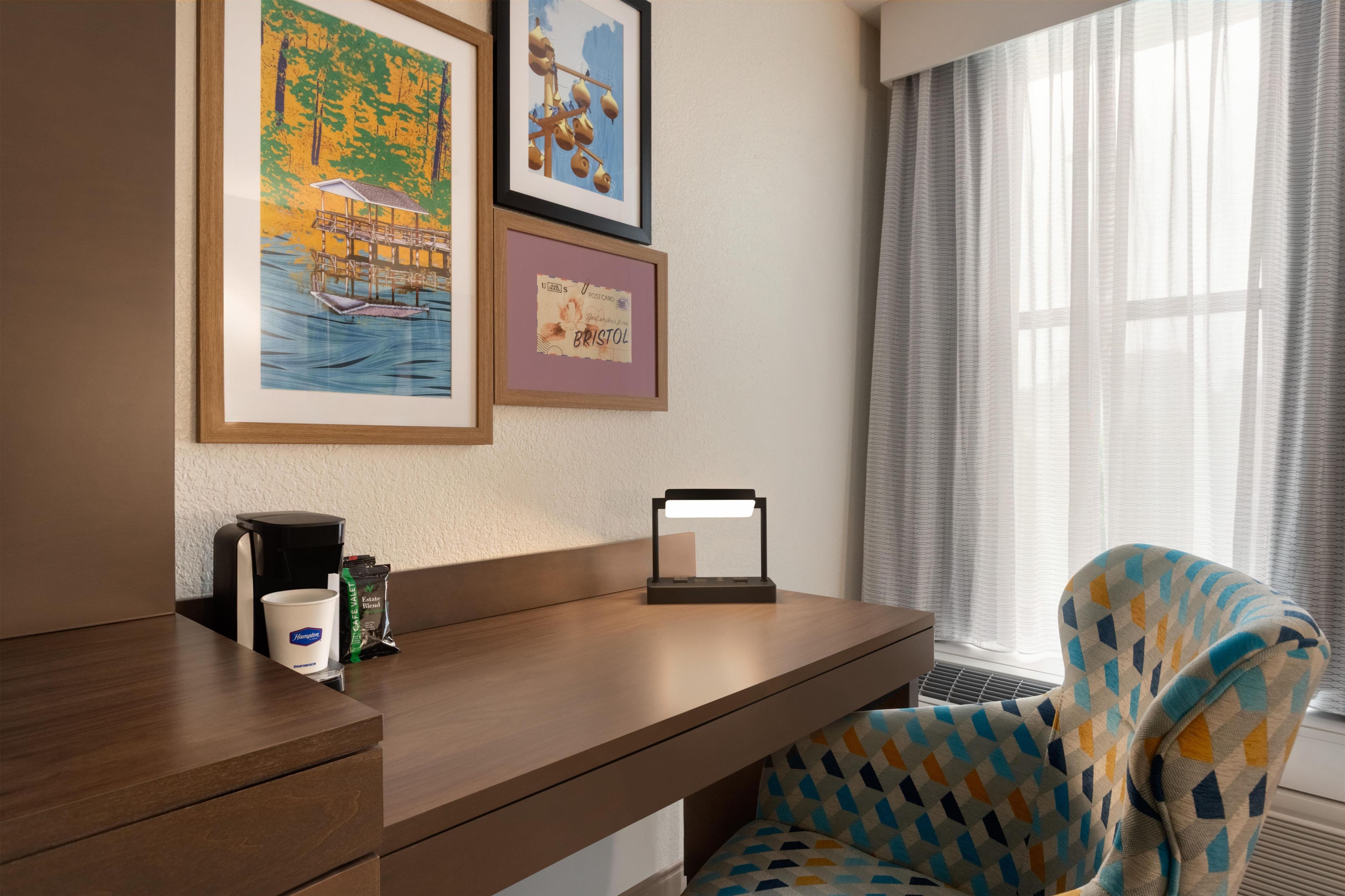 Habitación de hotel con un escritorio de madera, una silla con cojín estampado, cafetera y cuadros en la pared.
