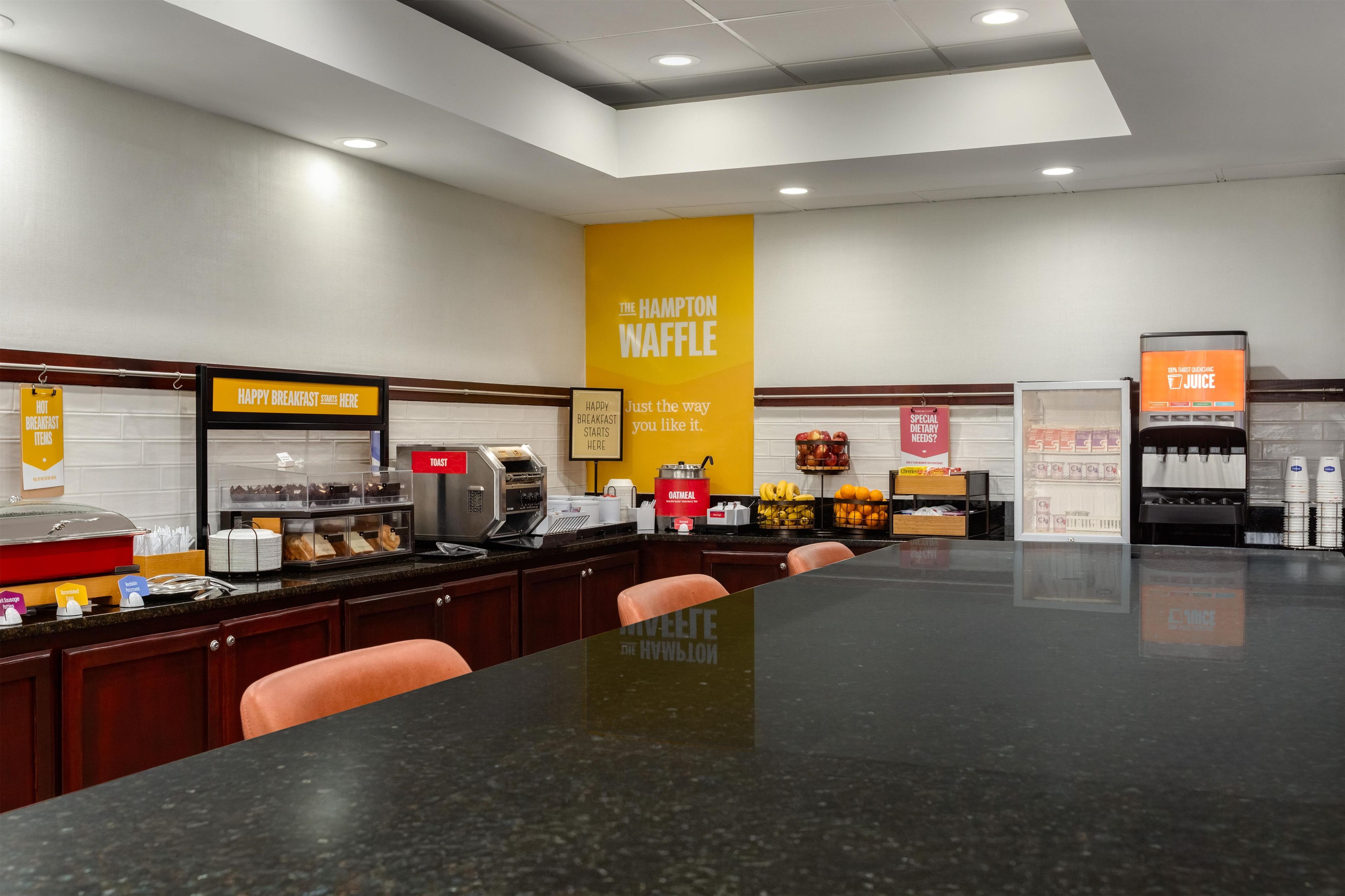 A breakfast area with a counter, seating, and various food service stations including a waffle station and juice dispenser.